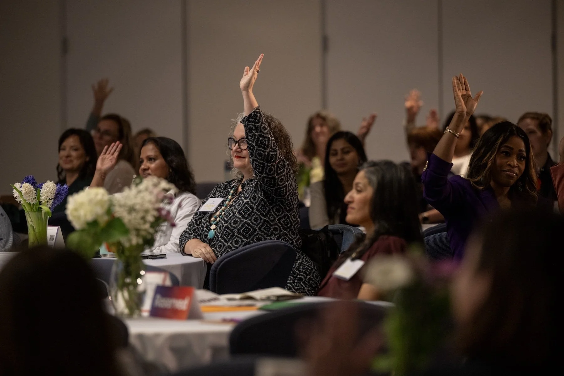 Women attending a conference, some raising their hands, sitting at round tables with flower centerpieces.