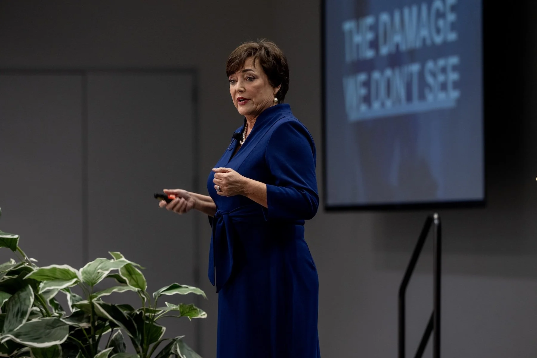 A woman in a blue dress giving a presentation in a conference room, with a large screen displaying text behind her.