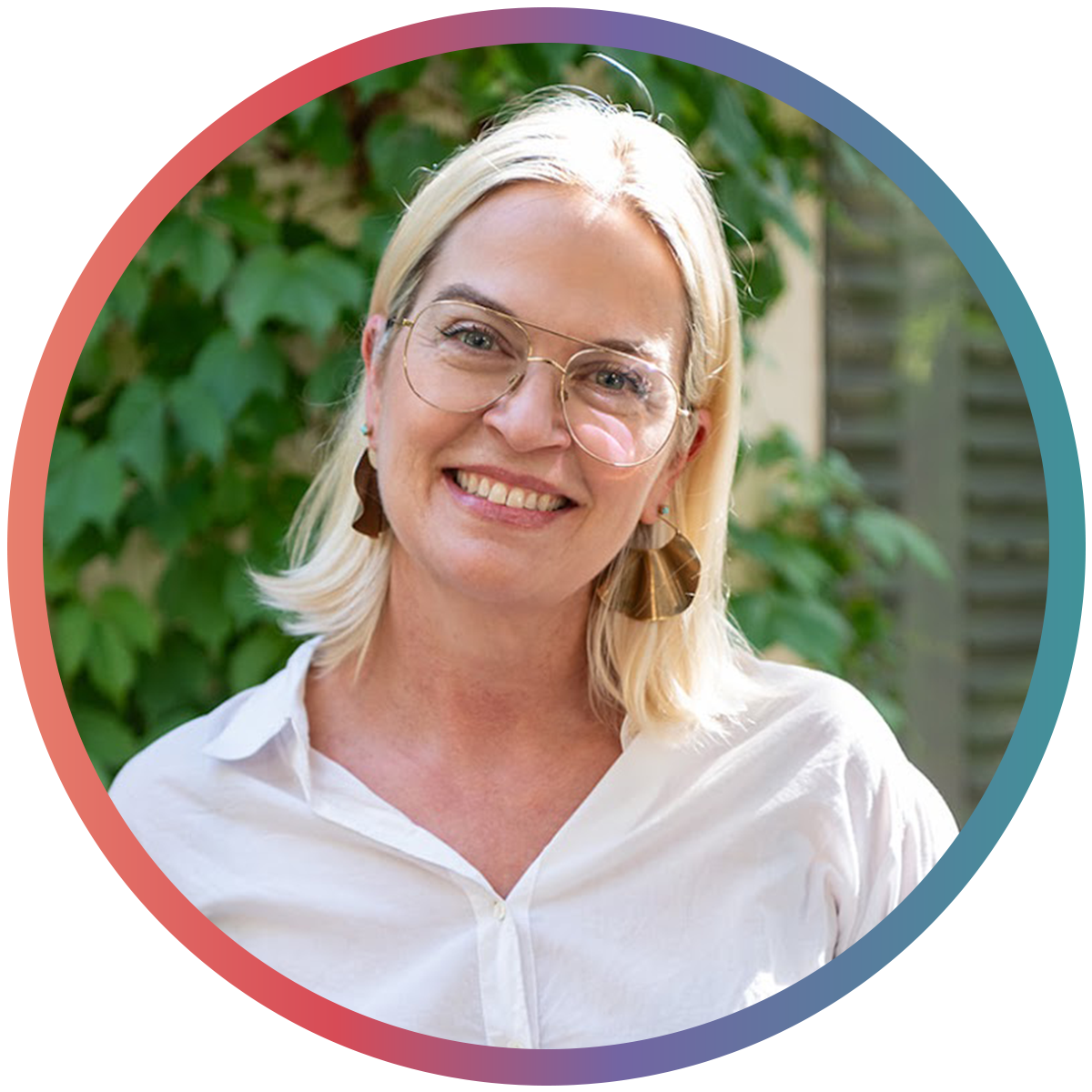 A smiling woman with blonde hair wearing glasses and earrings, standing outdoors with green foliage in the background.