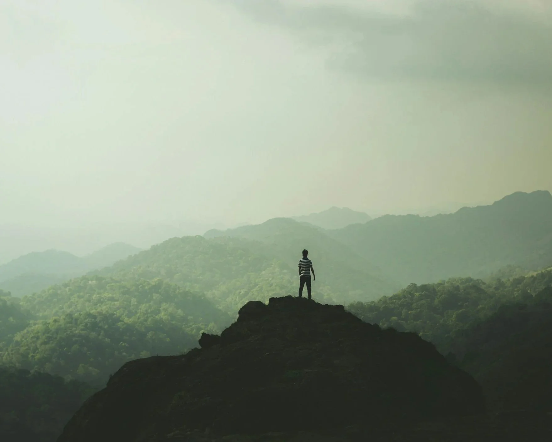 Eine Person steht auf einem Felsen und blickt auf hügelige, bewaldete Landschaften mit Bergen im Hintergrund, alles unter einem bewölkten Himmel.