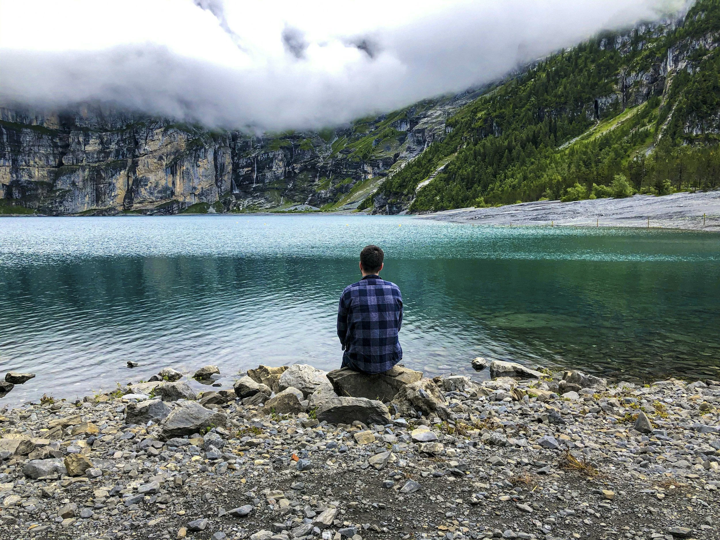 Ein Mann sitzt auf Steinen am Ufer eines blauen Sees, umgeben von Bergen und dichten Wäldern, bei bewölktem Himmel.