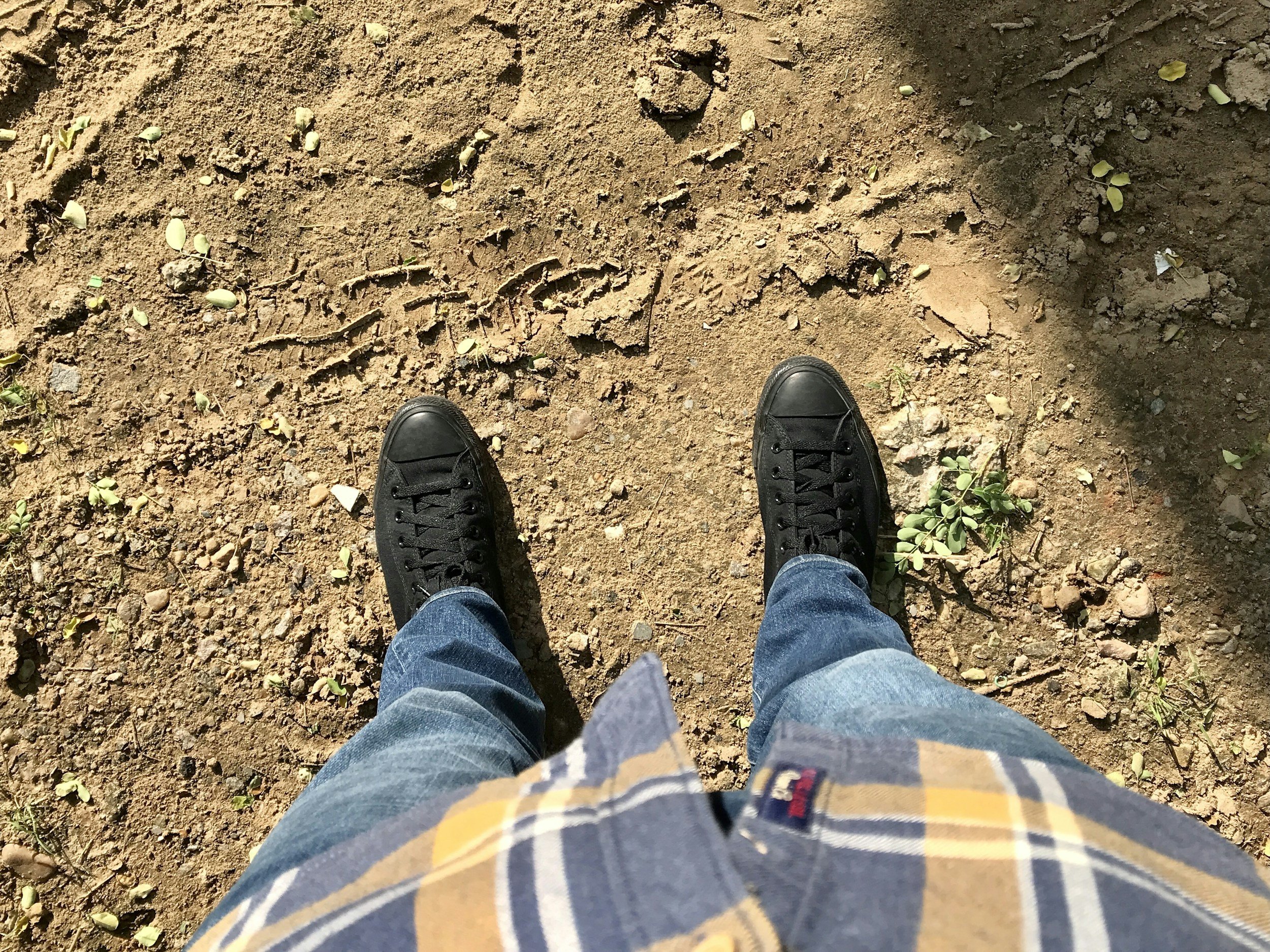 Perspektive eines stehenden Menschen mit schwarzen Schuhen, blauer Jeans und kariertem Hemd auf einem erdigen, befahrenen Boden mit kleinen Pflanzen und Fußspuren.