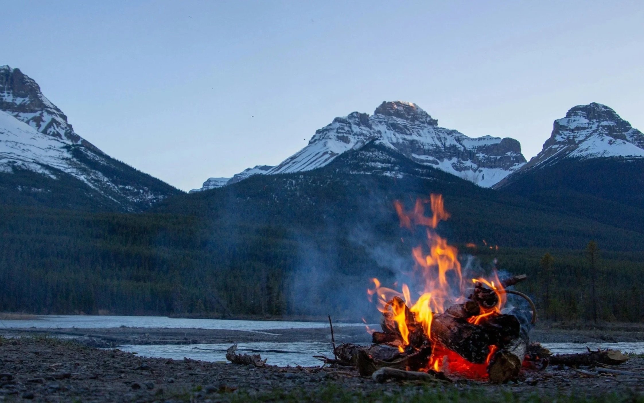 Ein Lagerfeuer brennt vor einer Bergkulisse mit schneebedeckten Gipfeln, in einer natürlichen, abgelegenen Gegend.