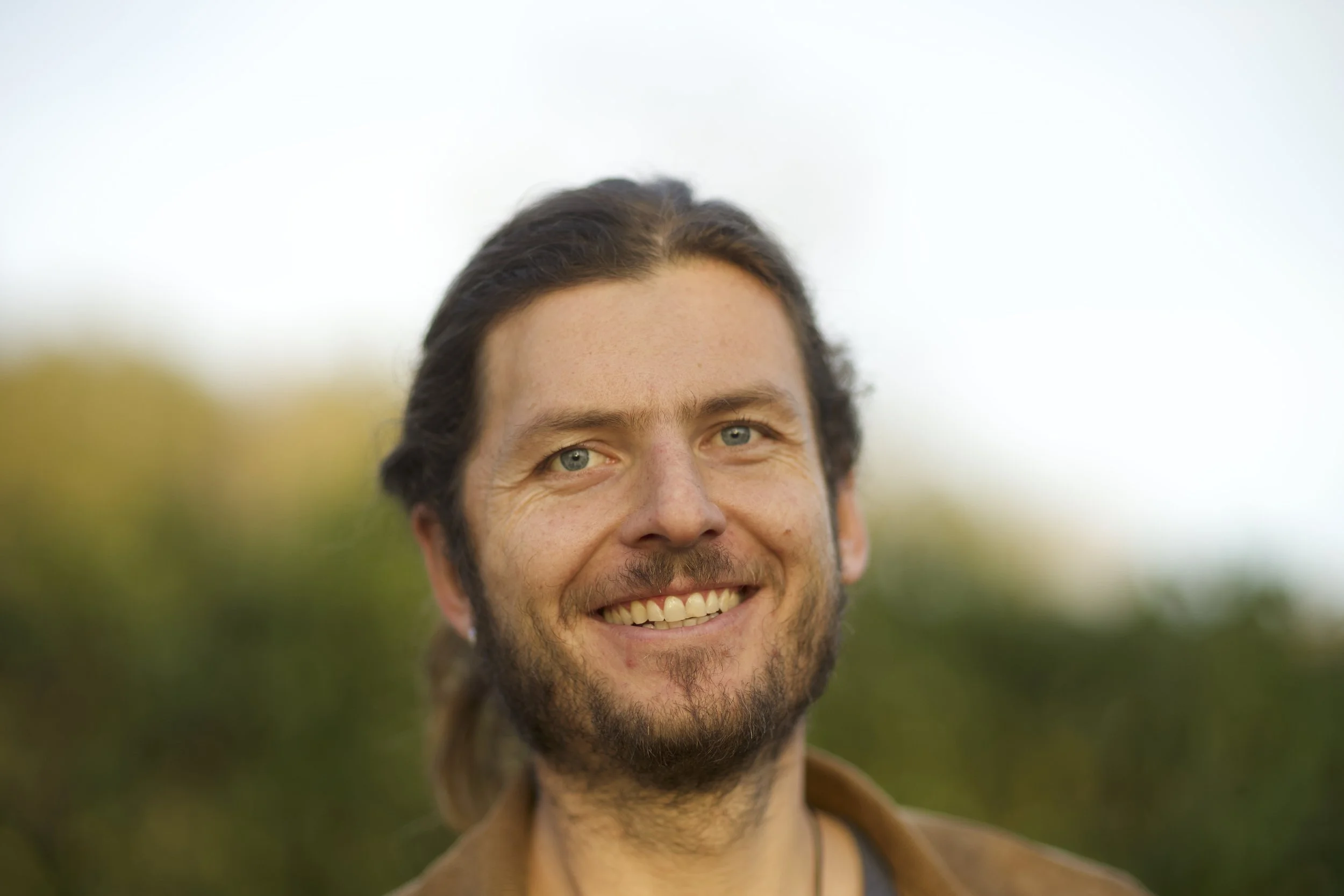 Ein lachender Mann mit dunklem Haar und Bart vor einem grünen, unscharfen Hintergrund, vermutlich draußen im Freien.