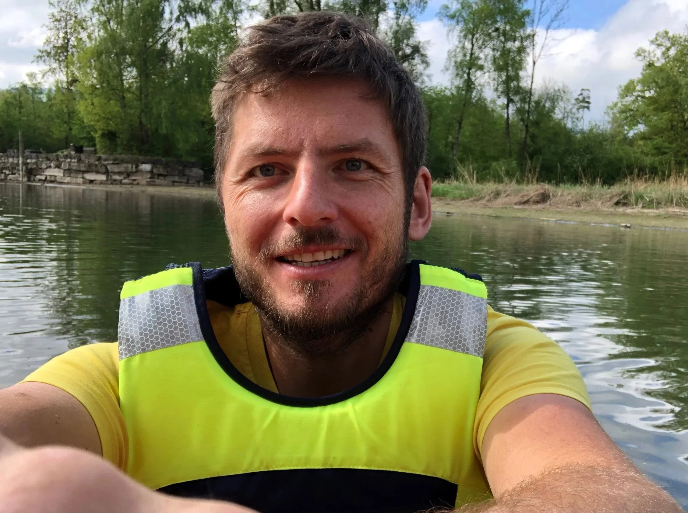 Ein lachender Mann mit braunem Haar und Bart, trägt einen neon-gelben Schwimmweste und ein gelbes T-Shirt, macht ein Selfie vor einer Wasserlandschaft mit Bäumen und Wolken im Himmel.