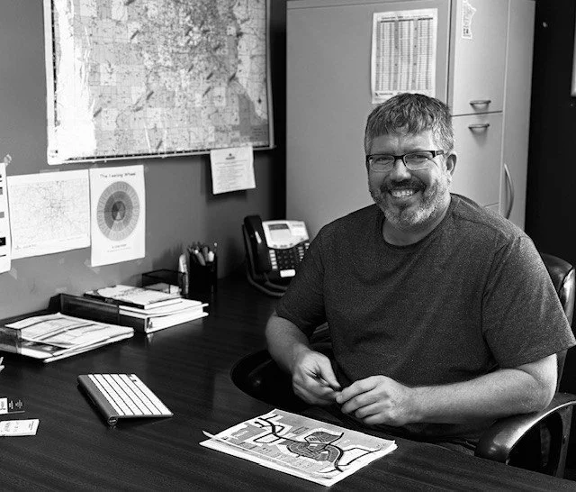 A man with glasses and a beard sitting at a desk in an office, smiling at the camera.