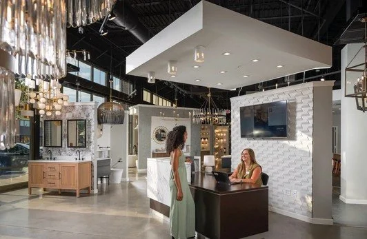 A modern house with wooden and white exterior walls in a mountainous landscape. The house has a large front window showing two people inside, with one visible silhouette and the other partially visible. The surrounding terrain is covered in yellowish grass and small shrubs, with mountains in the background.