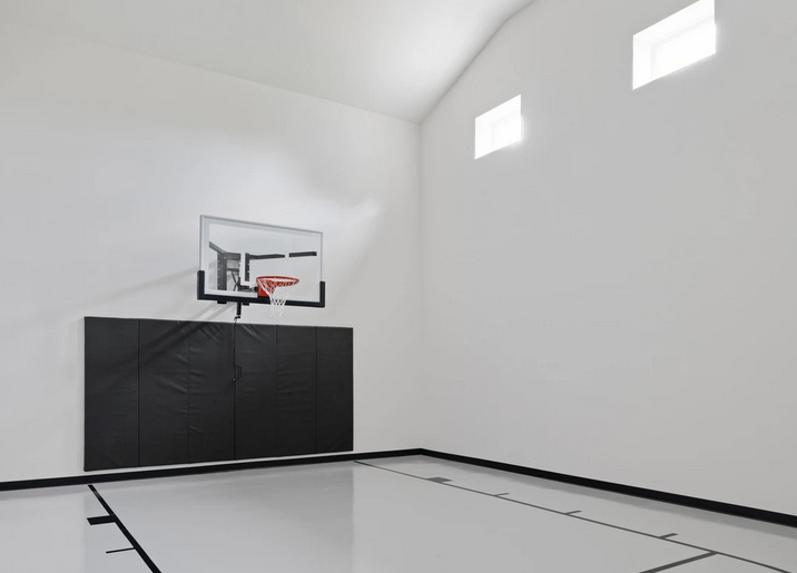 Empty indoor basketball court with a hoop mounted on the white wall, black padding below the hoop, and two small high windows allowing natural light.