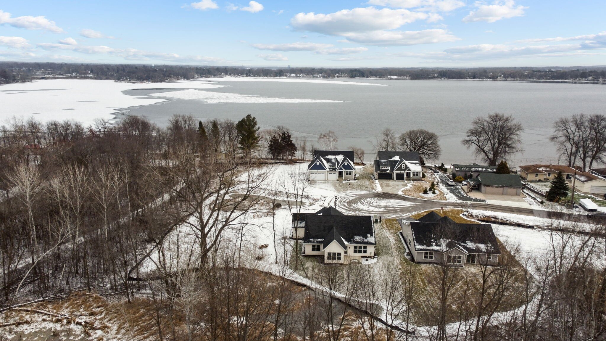 A lakeside residential area in winter with snow-covered ground, leafless trees, and houses. The lake is partially frozen with ice and snow.