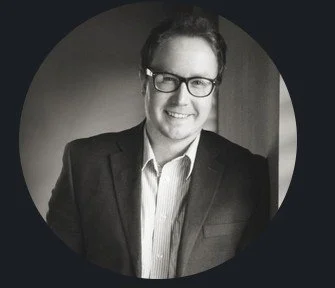 Black and white photo of a smiling man wearing glasses, a suit, and a collared shirt, indoors near a window.