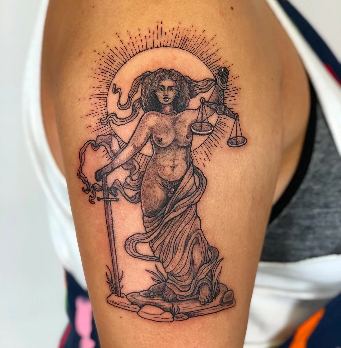 A tattoo of Lady Justice featuring a blindfold, holding scales and a sword, with intricate line work and shading, on a person's upper arm.