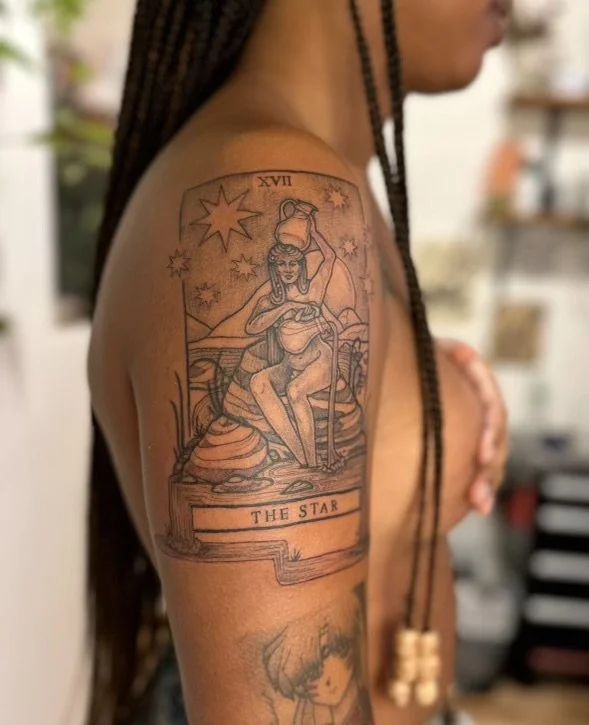 Tattoo on upper arm of a woman with long braided hair, sitting on a rock, holding a star-shaped object above her head. The tattoo features celestial elements and mountains in the background, with the words 'THE STAR' at the bottom.