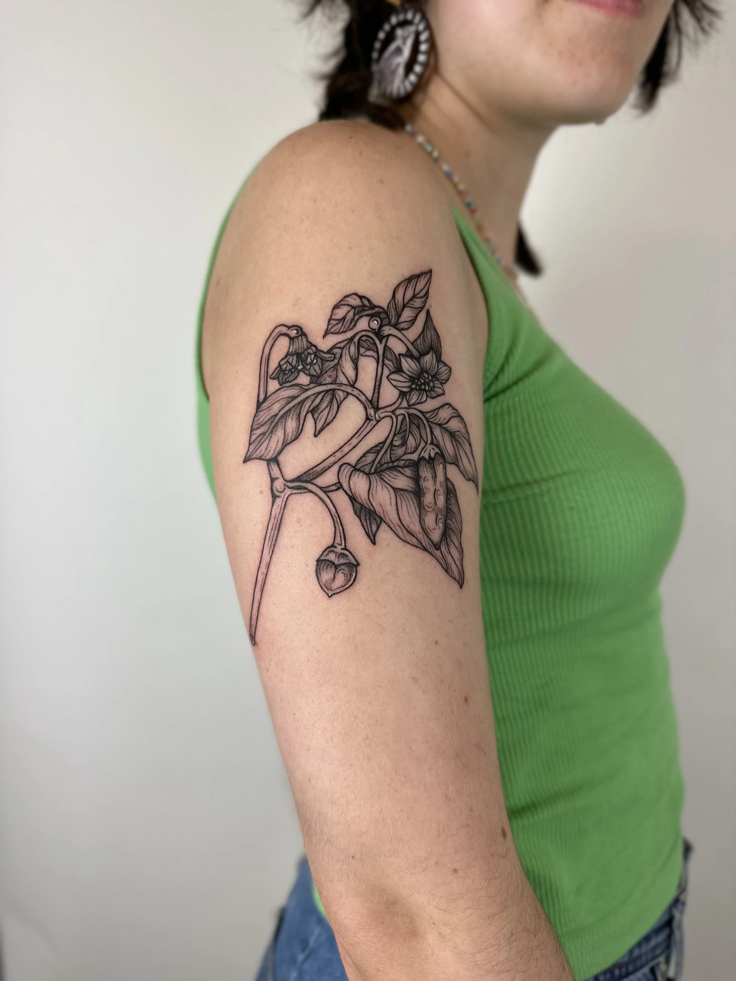A woman with a green tank top displaying a black line art tattoo of a plant with leaves, flowers, and seed pods on her upper arm.