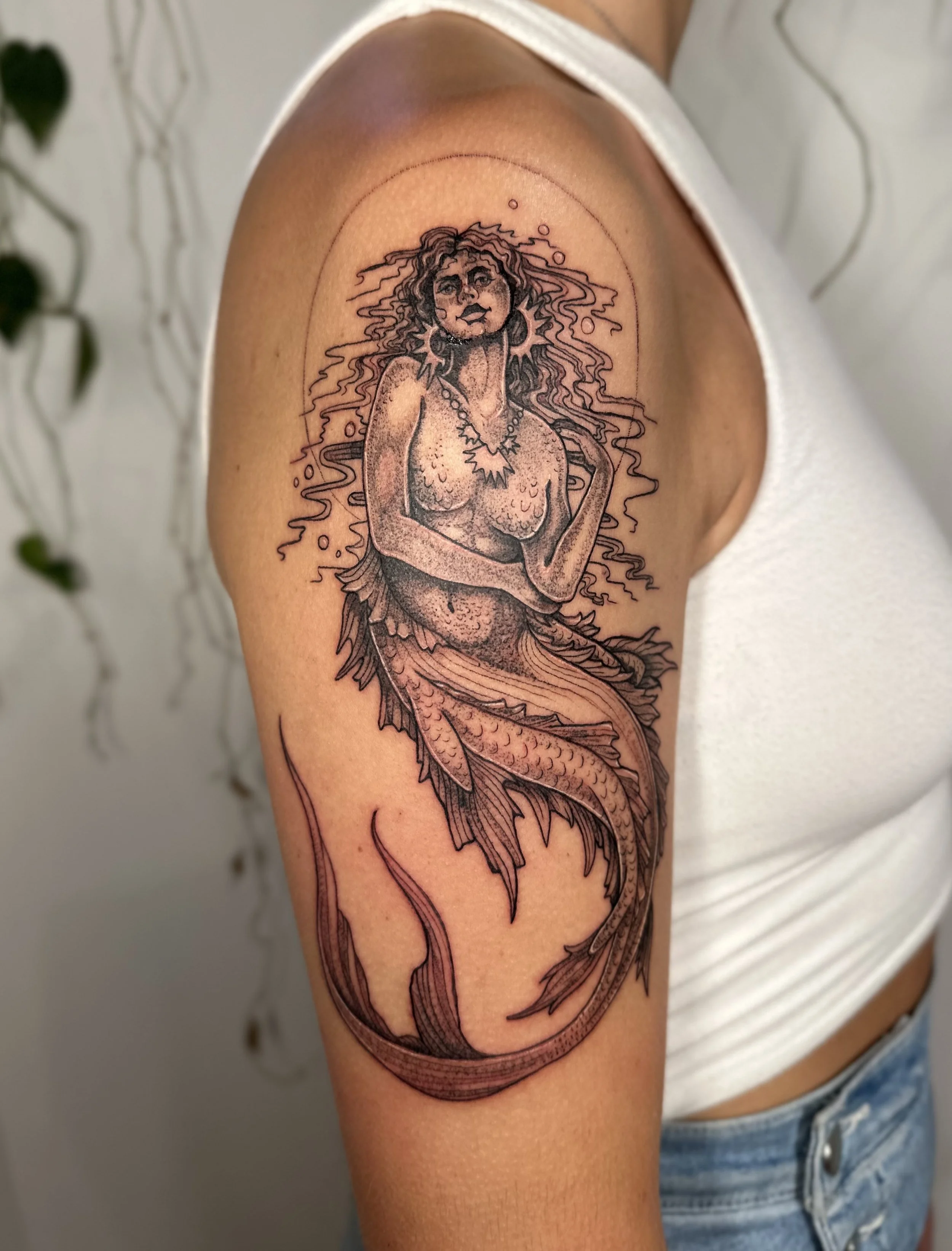 Tattoo of a mermaid with long, wavy hair, holding her chest, with a fish tail, on an upper arm.