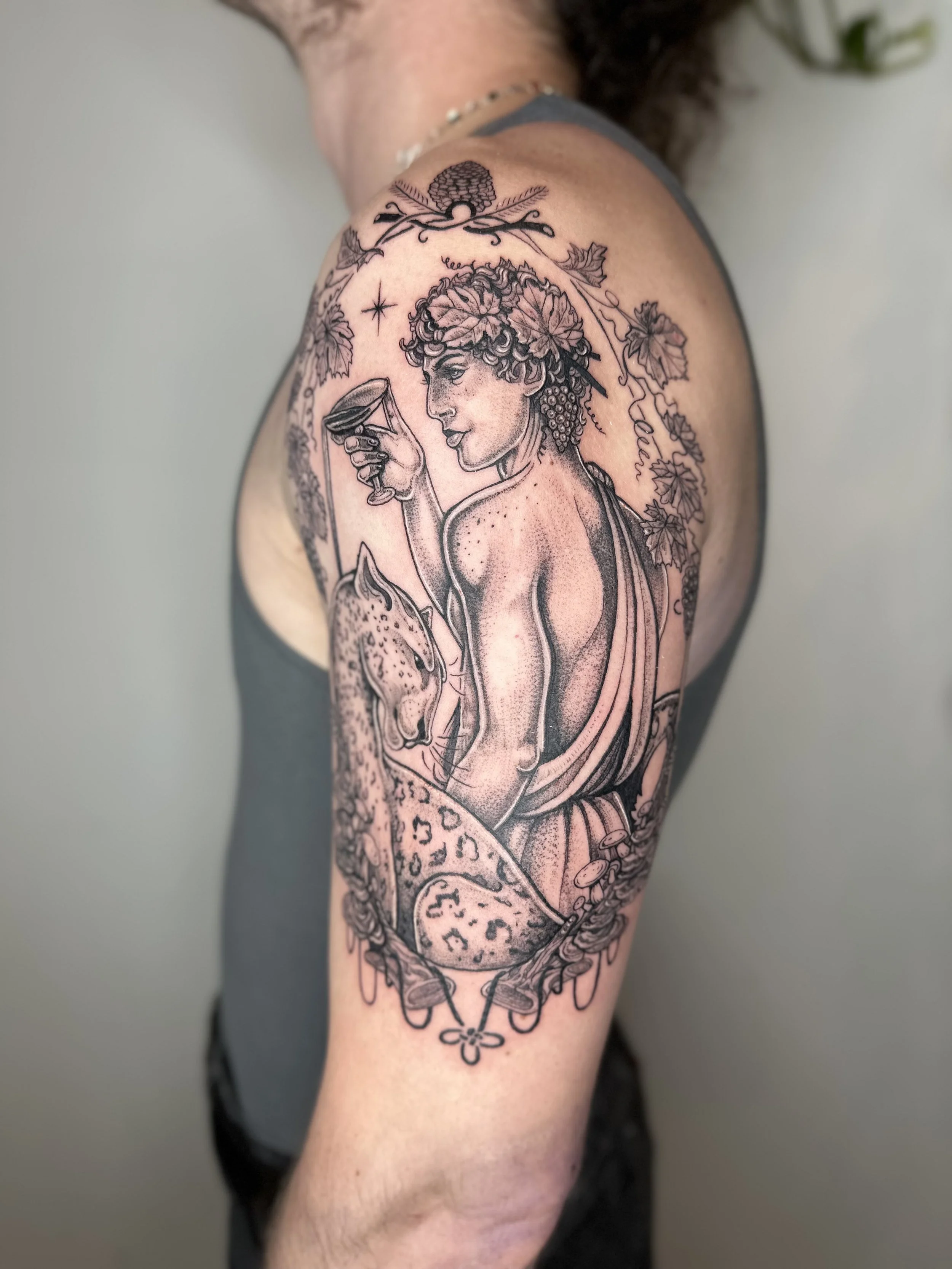 Tattoo of a man with curly hair, holding a glass, surrounded by grapes and vine, detailed with leaves and decorative elements.