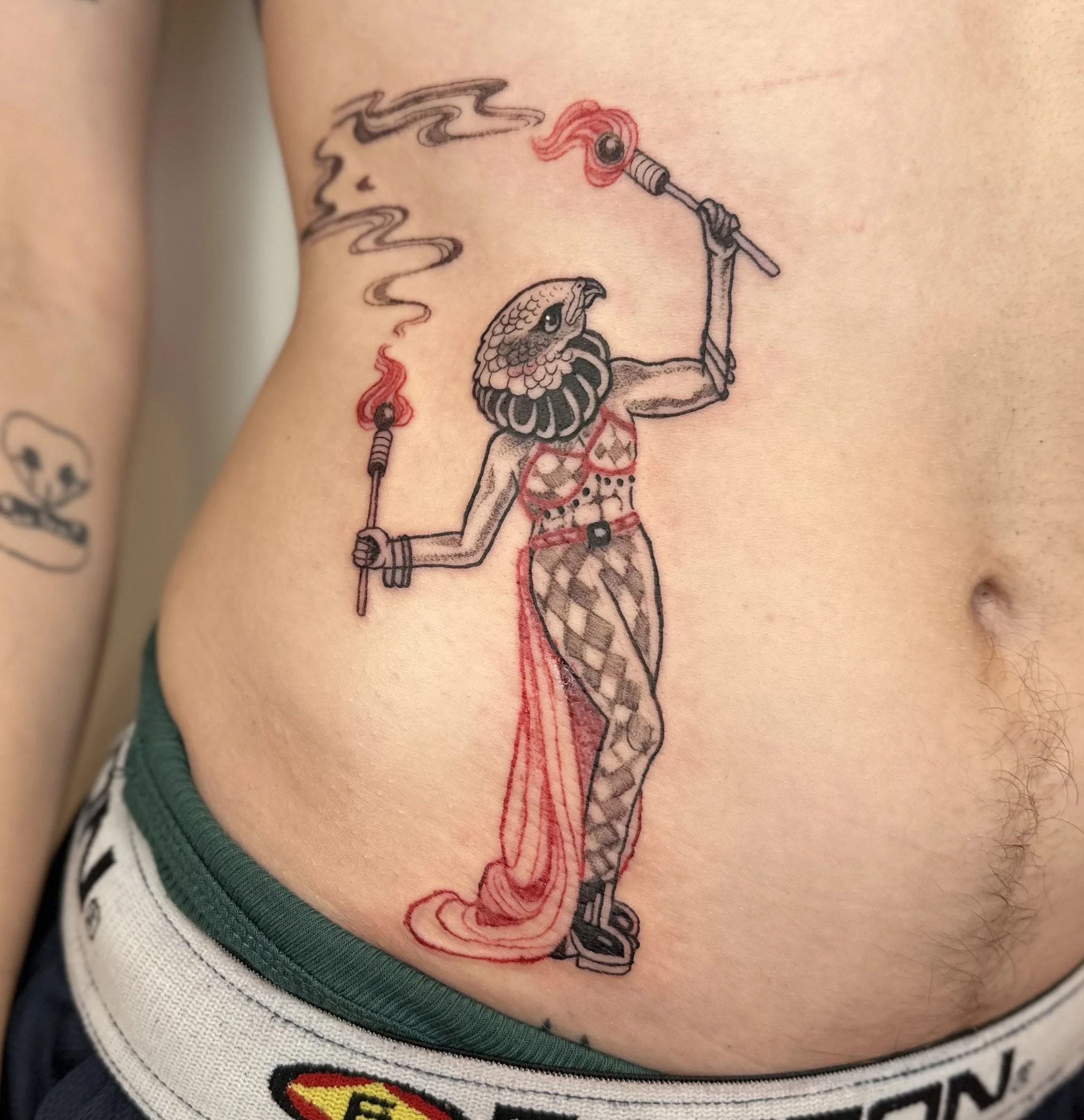 Colorful tattoo of a woman with a bird face holding torches with flames on the chest and legs, on the person's side.