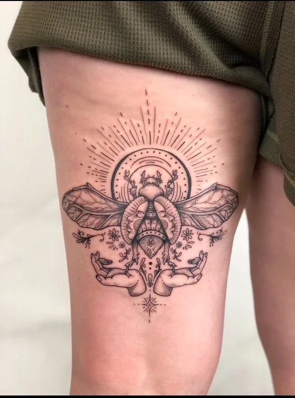 A detailed black ink tattoo of a moth with outstretched wings, decorated with floral and ornamental elements, centered on the upper thigh, with radiating lines and halo-like circle around its head.