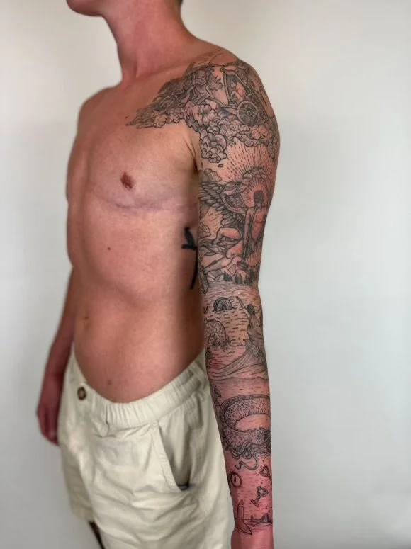 A shirtless man with tan skin and beige shorts, showcasing a detailed black and gray sleeve tattoo that features clouds, mountains, a sun, and various natural elements, standing against a plain white background.