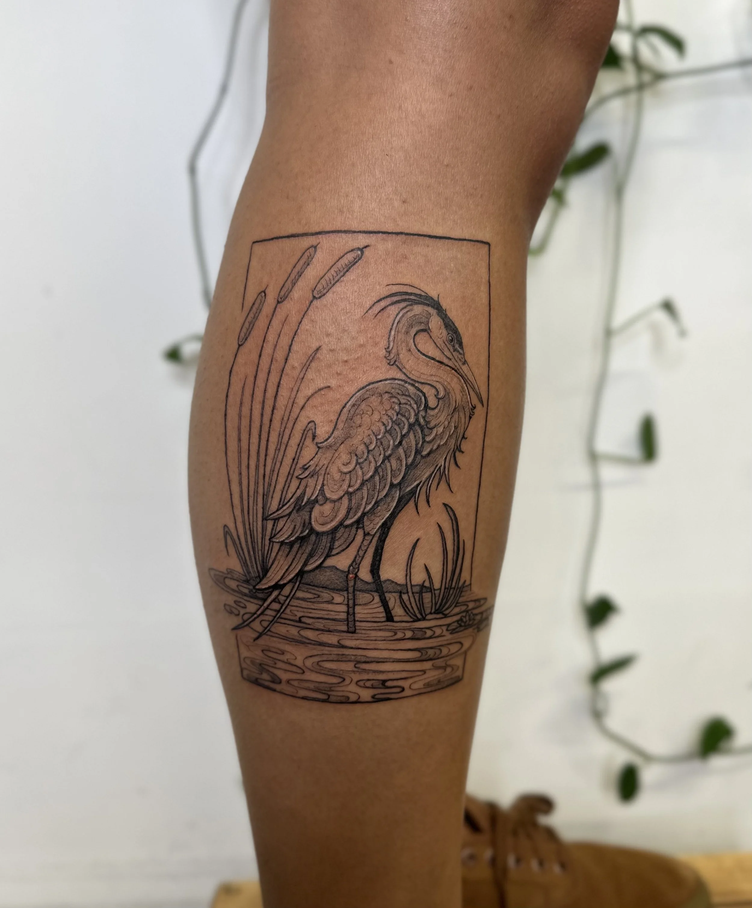 Line art tattoo of a heron standing in water, enclosed in a rectangular frame, on a person's thigh.