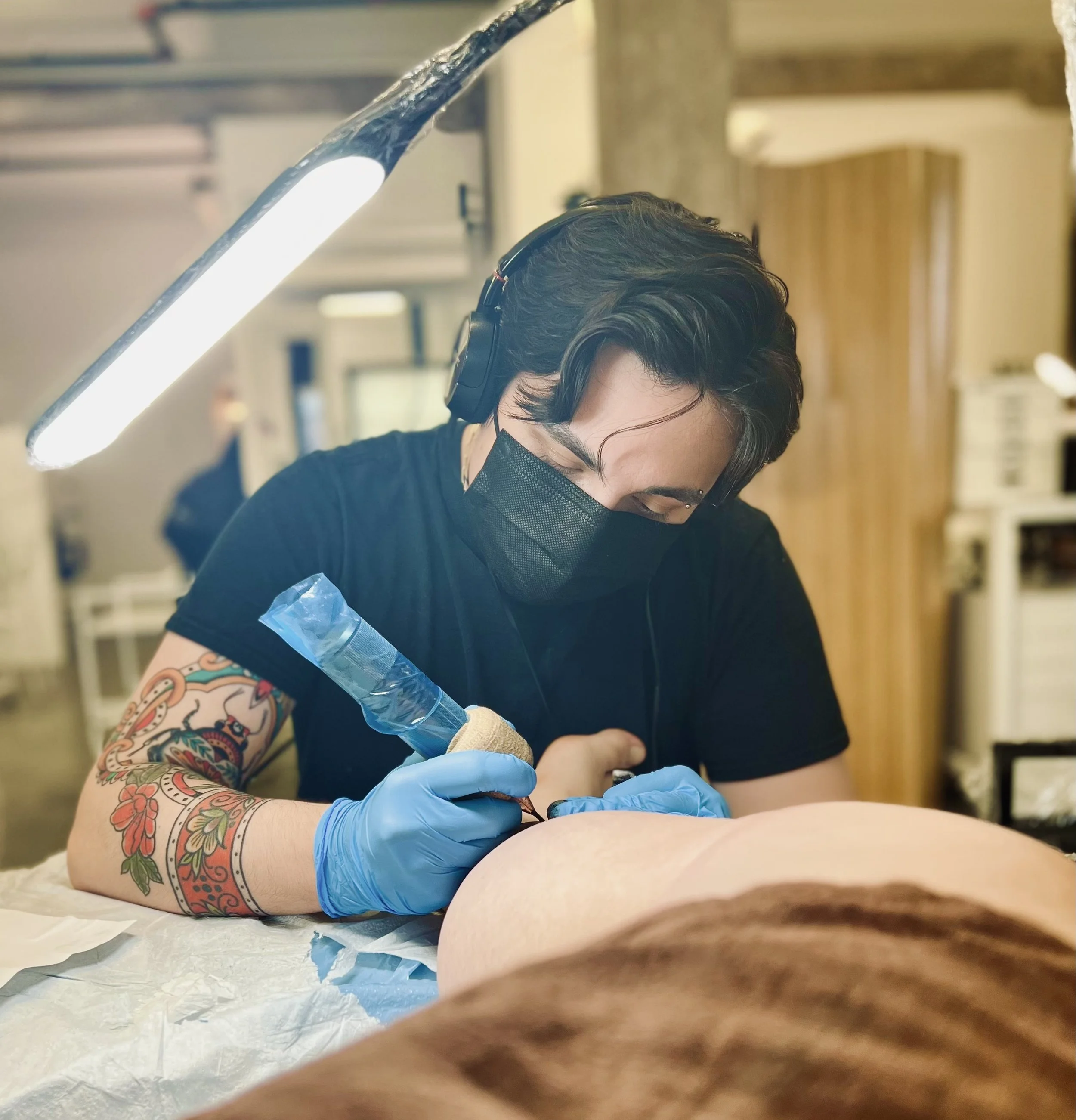 Tattooed artist wearing a black mask, black shirt, and gloves, tattooing a person's arm in a tattoo studio.
