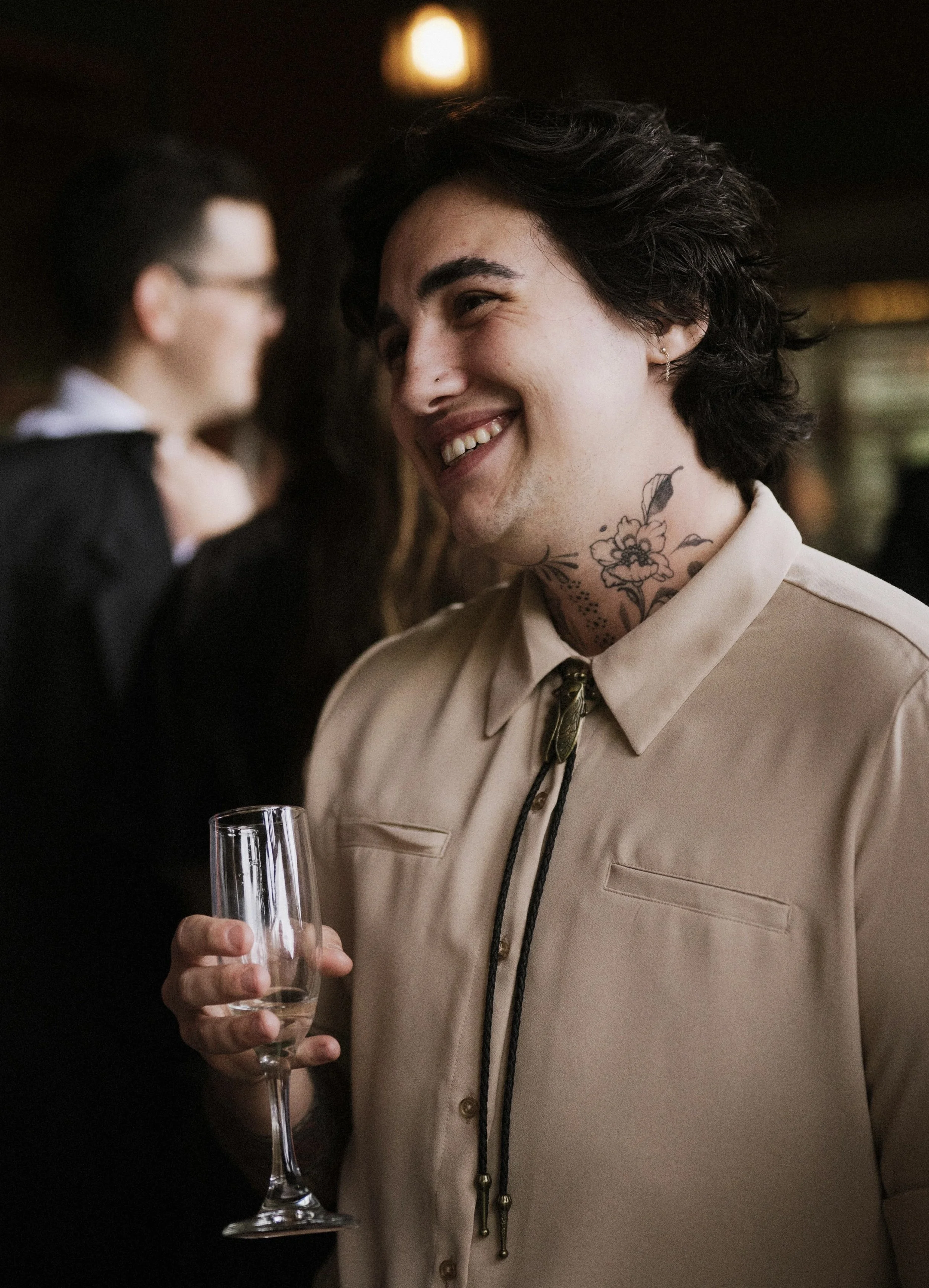 A young man with dark, curly hair, tattoos on his neck, and wearing a beige shirt holding a glass of champagne, smiling at an event or party.