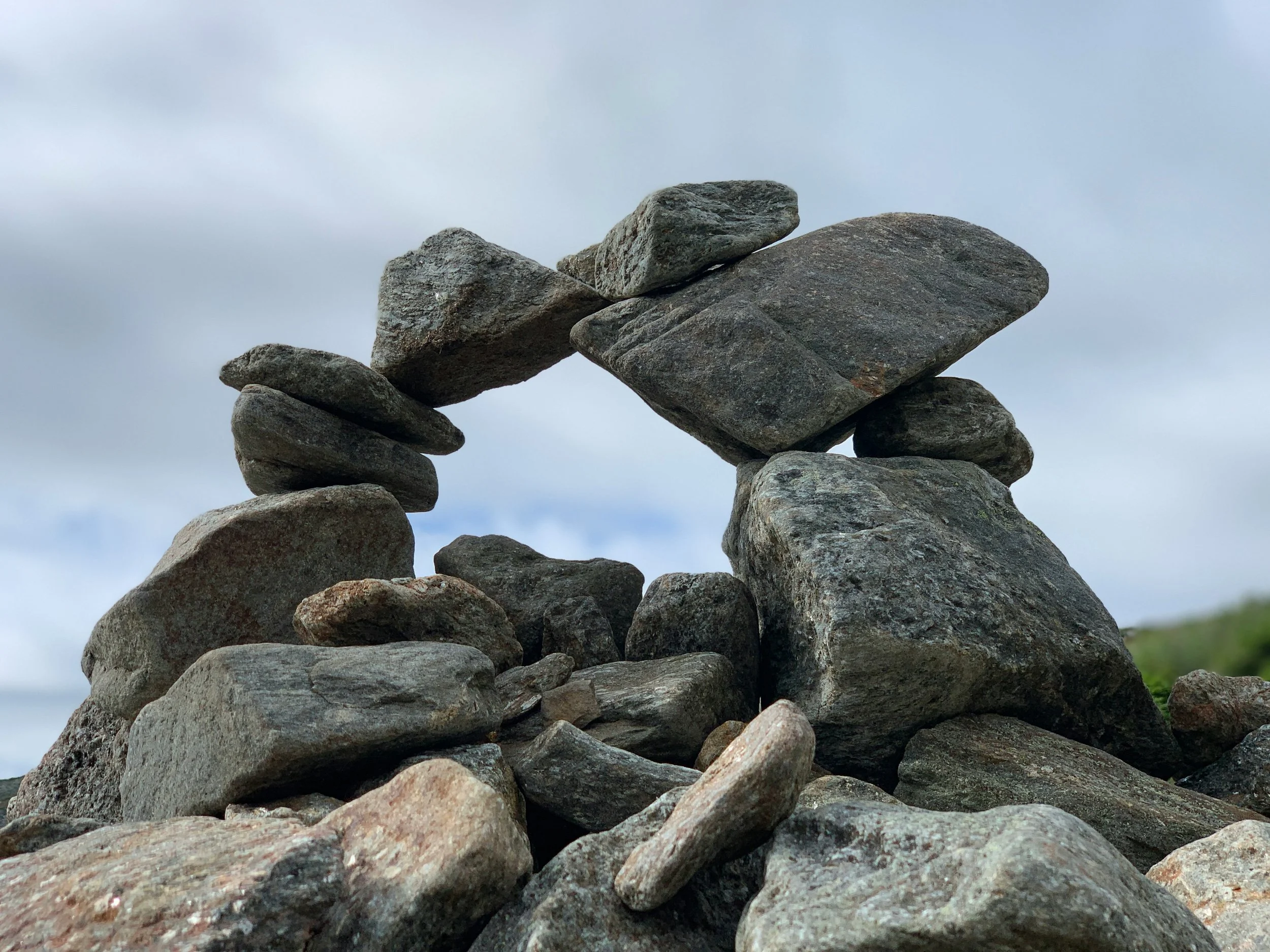 Stacked rocks forming a natural arch during daytime with cloudy sky and some greenery in the background.