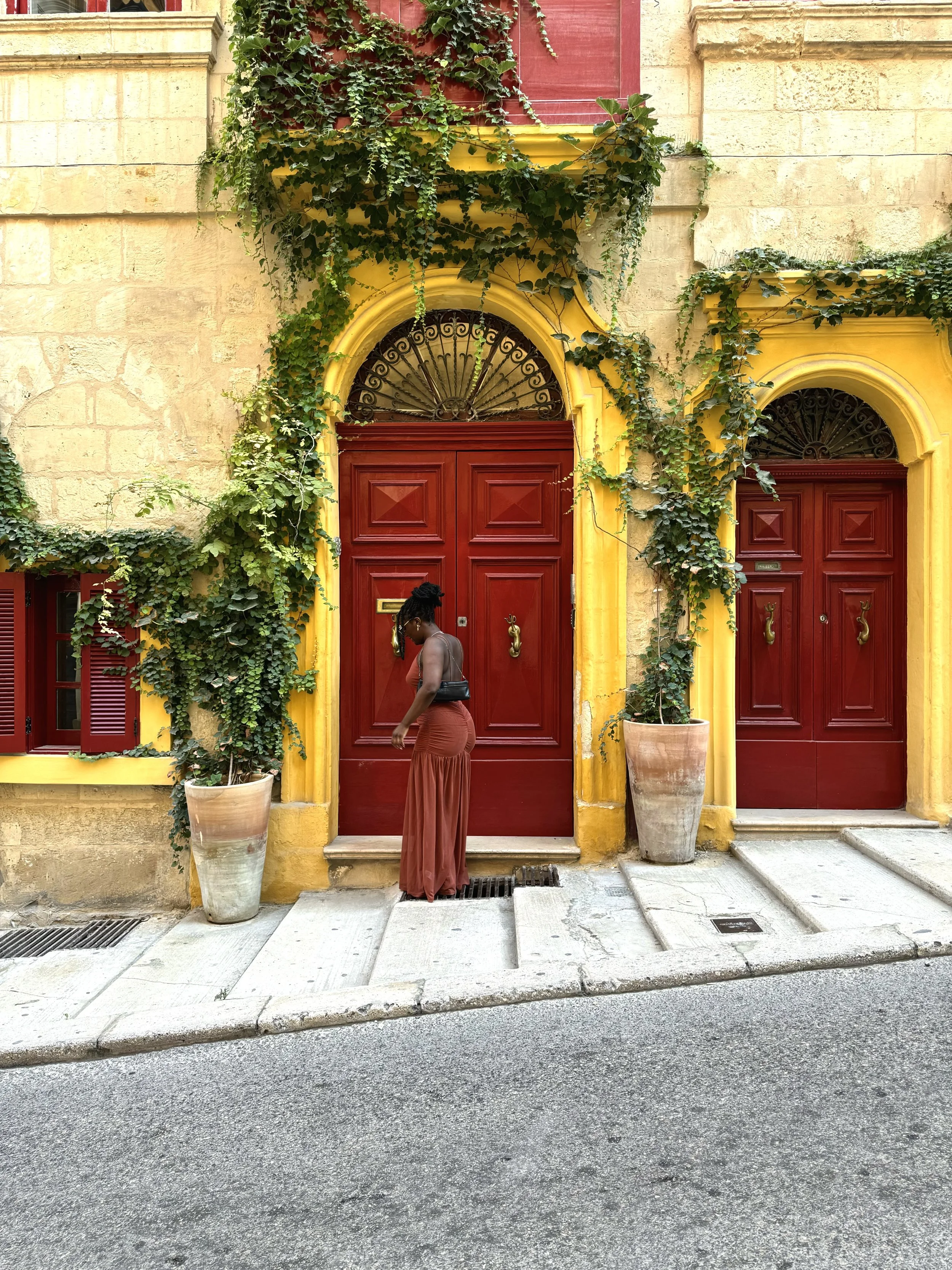 Personne marchant devant une façade colorée avec deux portes rouges, entourée de végétation verte et de pots de fleurs, murs en pierre beige.