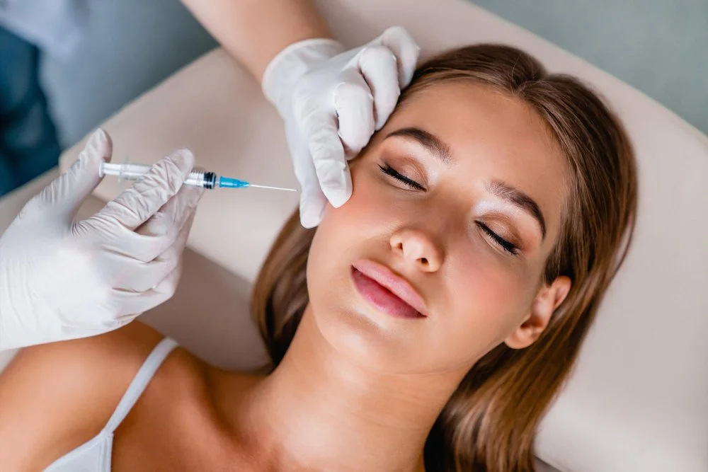 A woman is lying down with her eyes closed while a medical professional administers a cosmetic treatment with a syringe to her forehead.