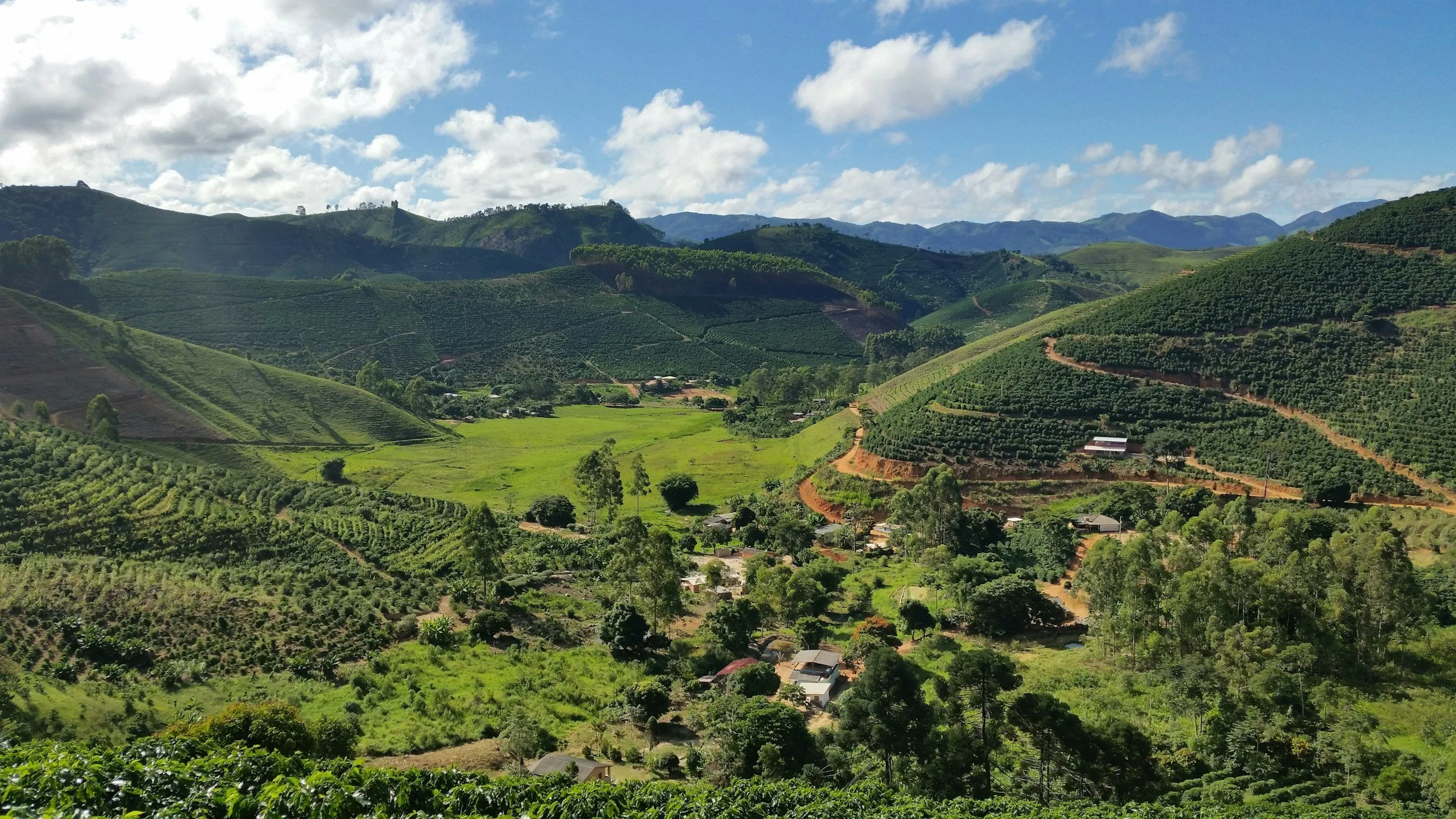 Green rolling hills and mountains with scattered houses, trees, and agricultural fields under a partly cloudy sky.