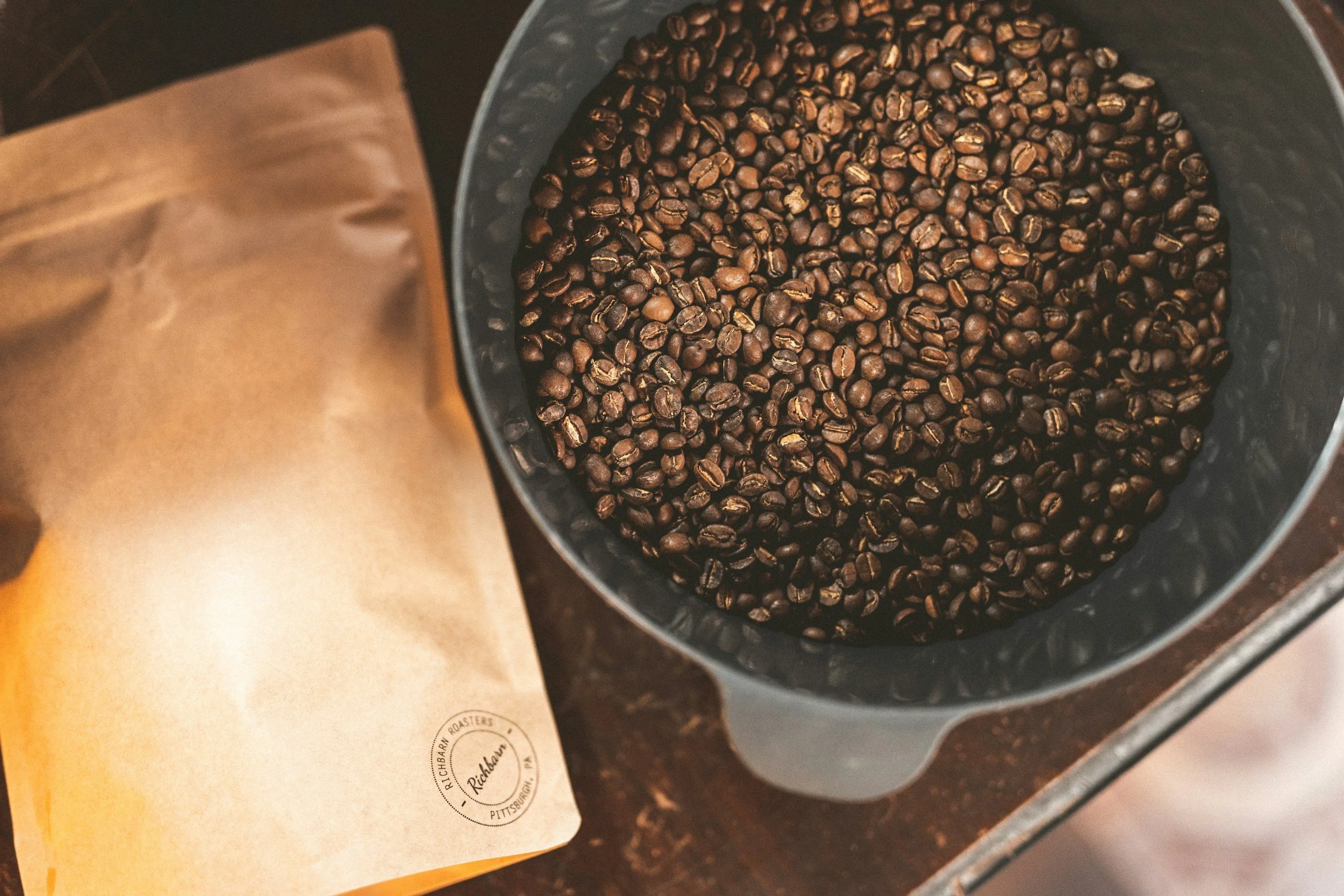 A container filled with roasted coffee beans next to a brown paper bag labeled "Richland" and "Pittsburgh".