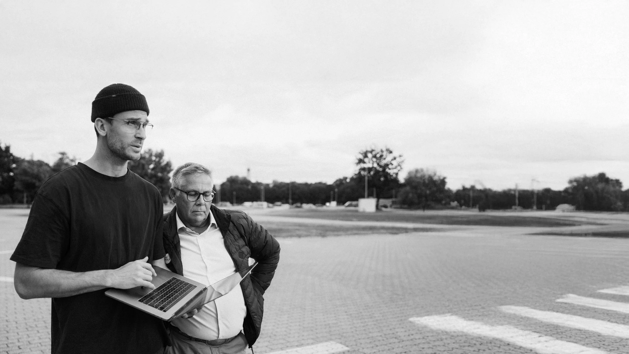Zwei Männer stehen auf einem Parkplatz, der mit Pflastersteinen gepflastert ist, und betrachten ein Laptop. Der jüngere Mann trägt eine schwarze Kappe, eine schwarze T-Shirt und eine Brille, während der ältere Mann eine weiße Bluse, eine schwarze Jacke und ebenfalls eine Brille trägt. Im Hintergrund sind Bäume, ein wenige PKWs und eine offene Landschaft sichtbar.
