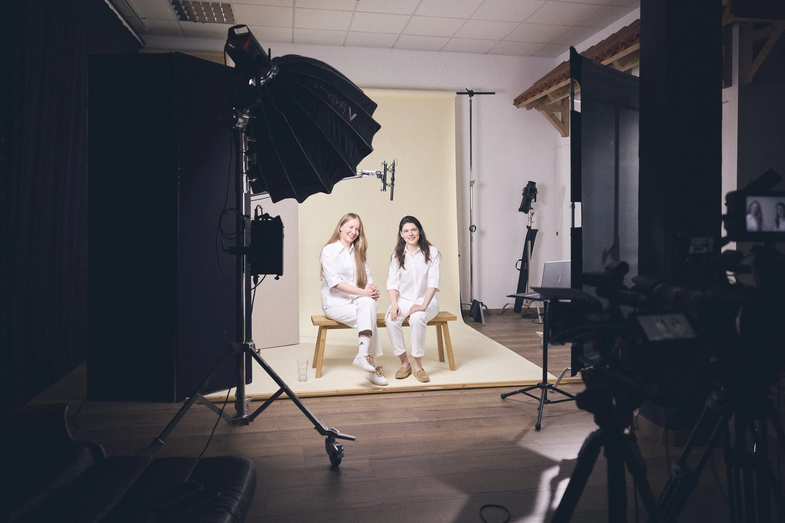 Zwei Frauen sitzen auf einer Bank vor einem weißen Hintergrund im Fotostudio, umgeben von professioneller Fotografie- und Videotechnik.