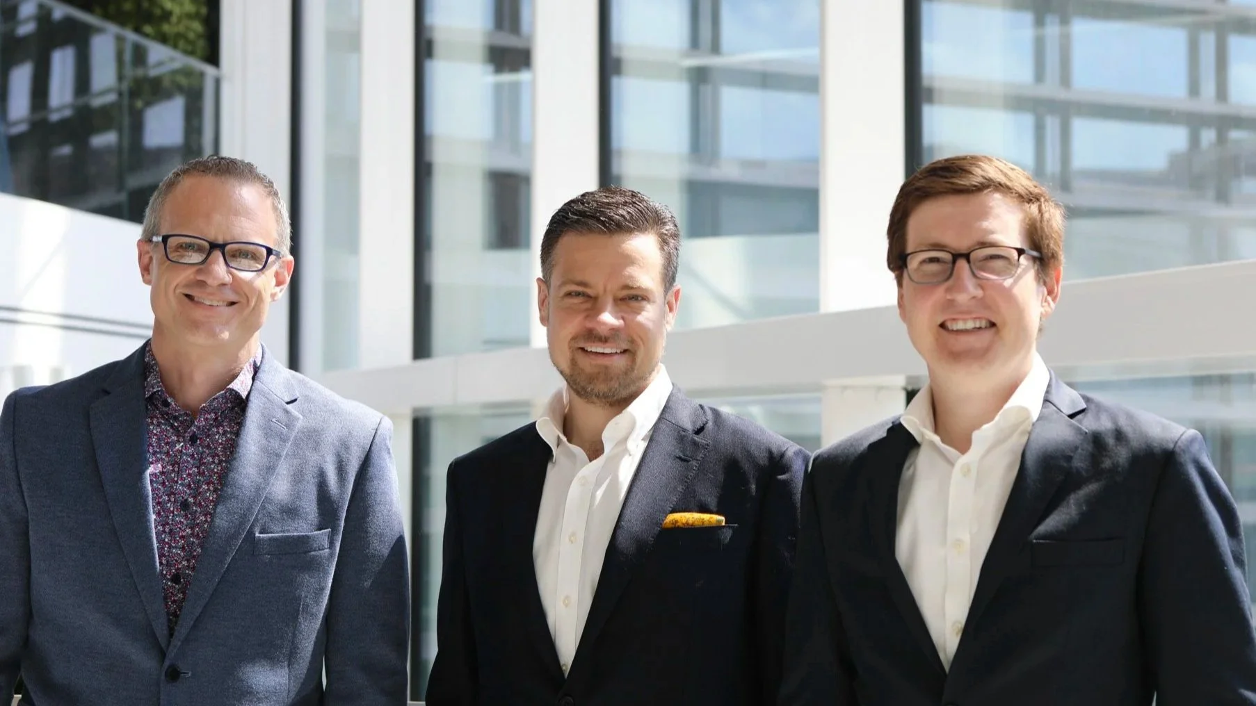Three happy men in business suits standing outside in front of a modern glass building.