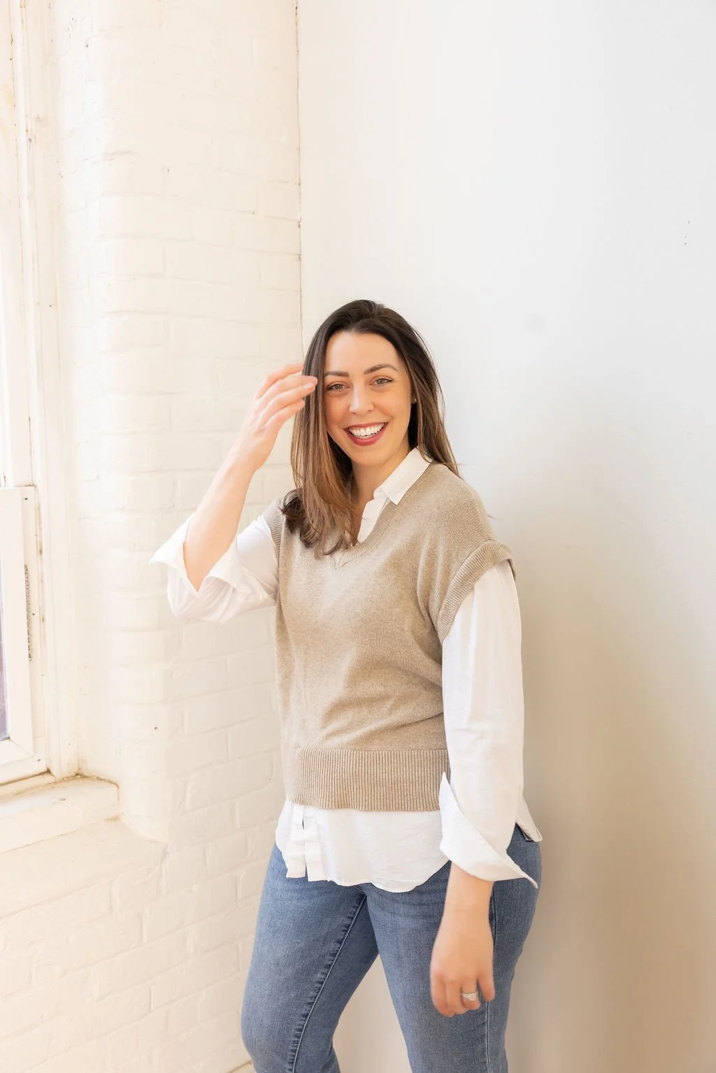 A woman smiling and touching her hair, standing near a white brick wall with a window.
