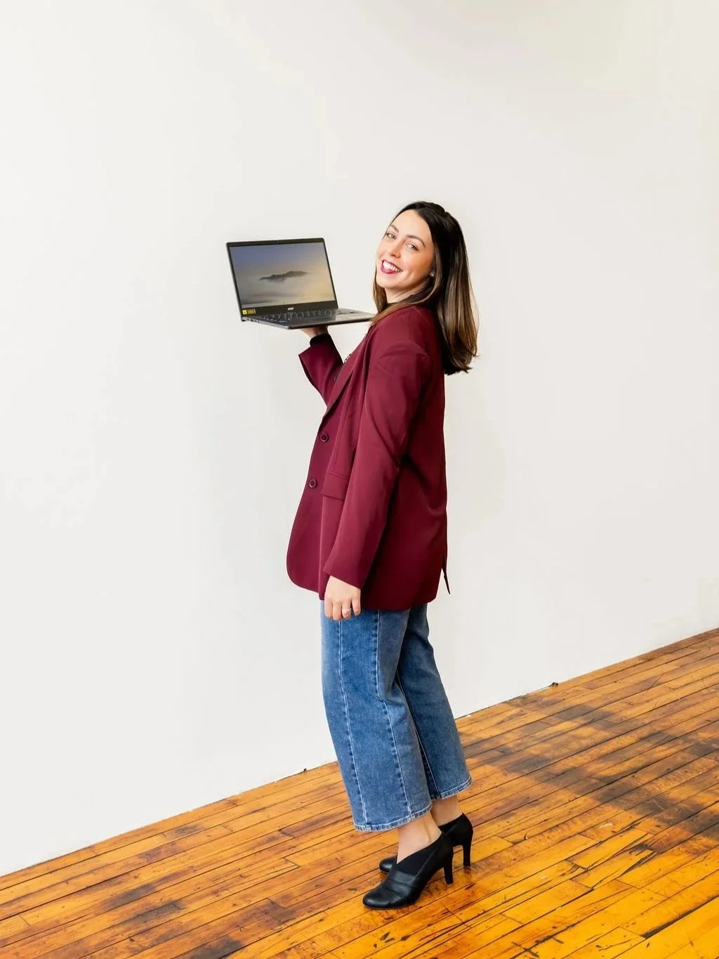 Kelly Martins wearing a burgundy blazer holding a laptop and looking over her shoulder at the camera, representing brand alignment, website strategy, and visibility challenges for service-based business owners.