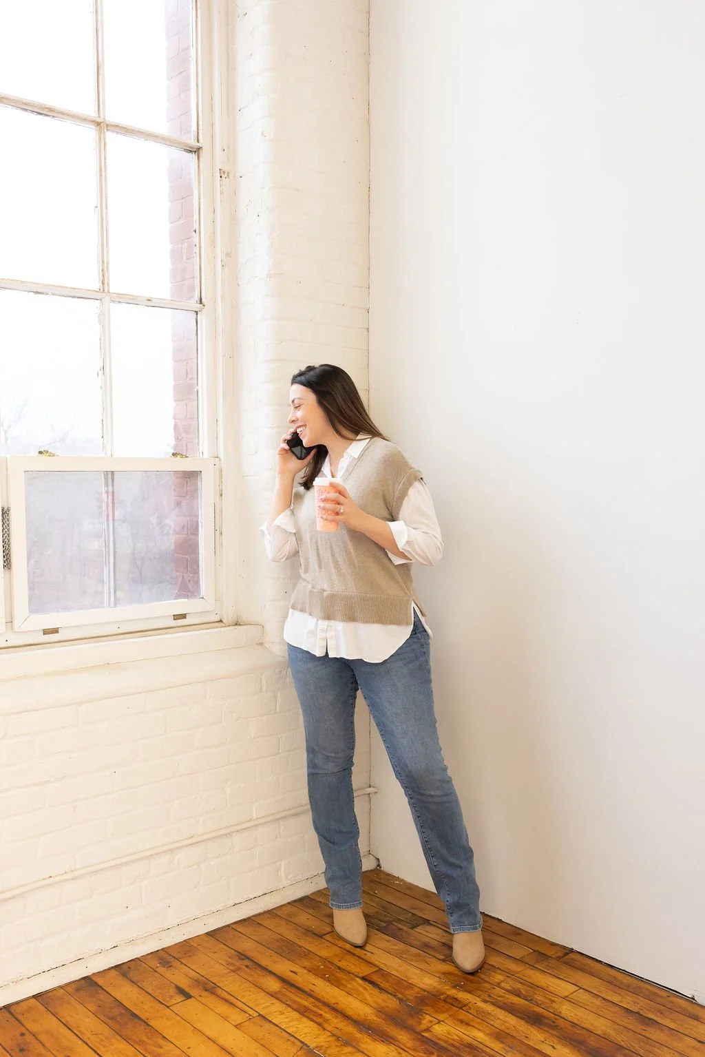 Woman smiling and talking on the phone while holding a coffee cup in a bright room with large windows and wooden floor.