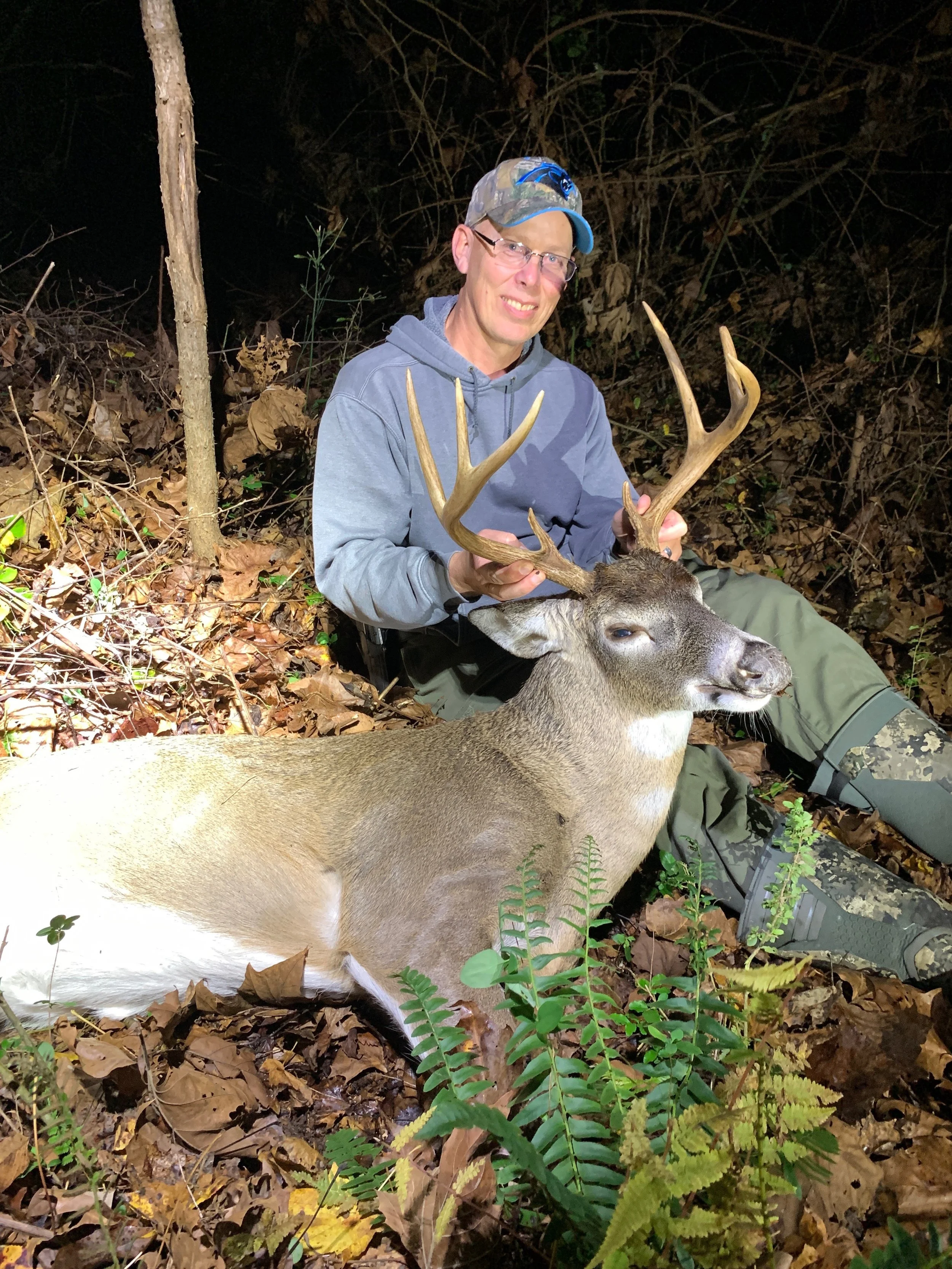 A man in outdoor gear sitting on the ground at night, holding the antlers of a large buck lying on leaves and ferns, in a wooded area.