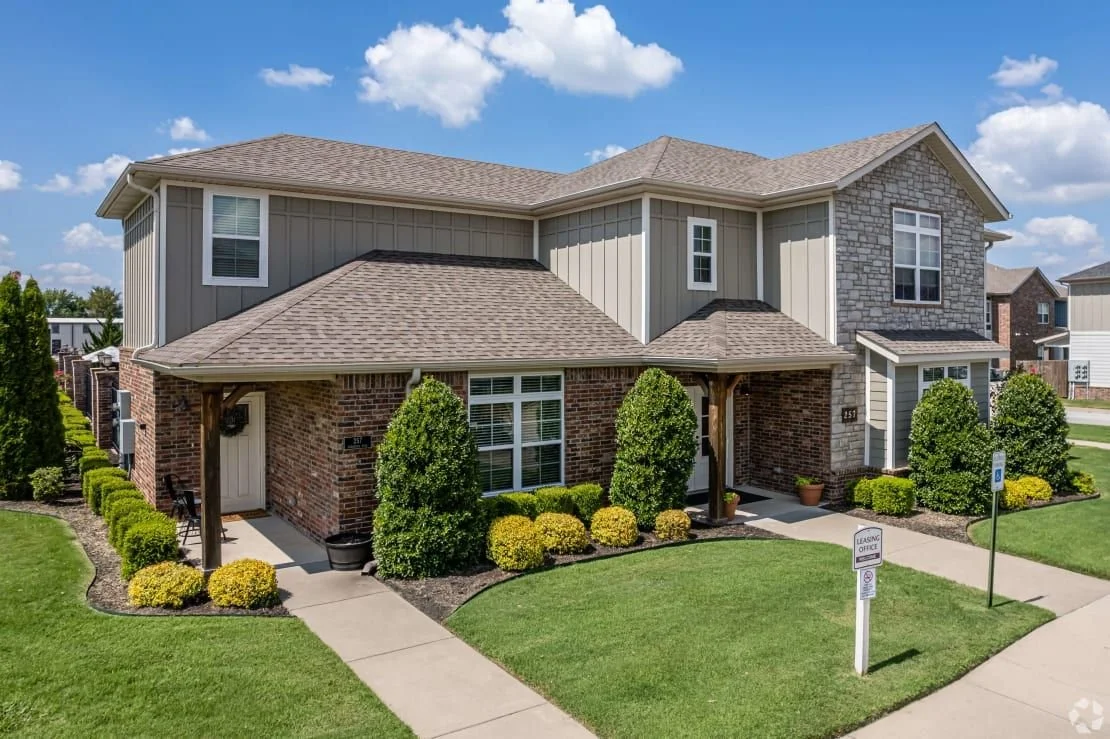 A two-story residential house with brick and siding exterior, surrounded by a well-maintained lawn and shrubbery, under a partly cloudy sky.