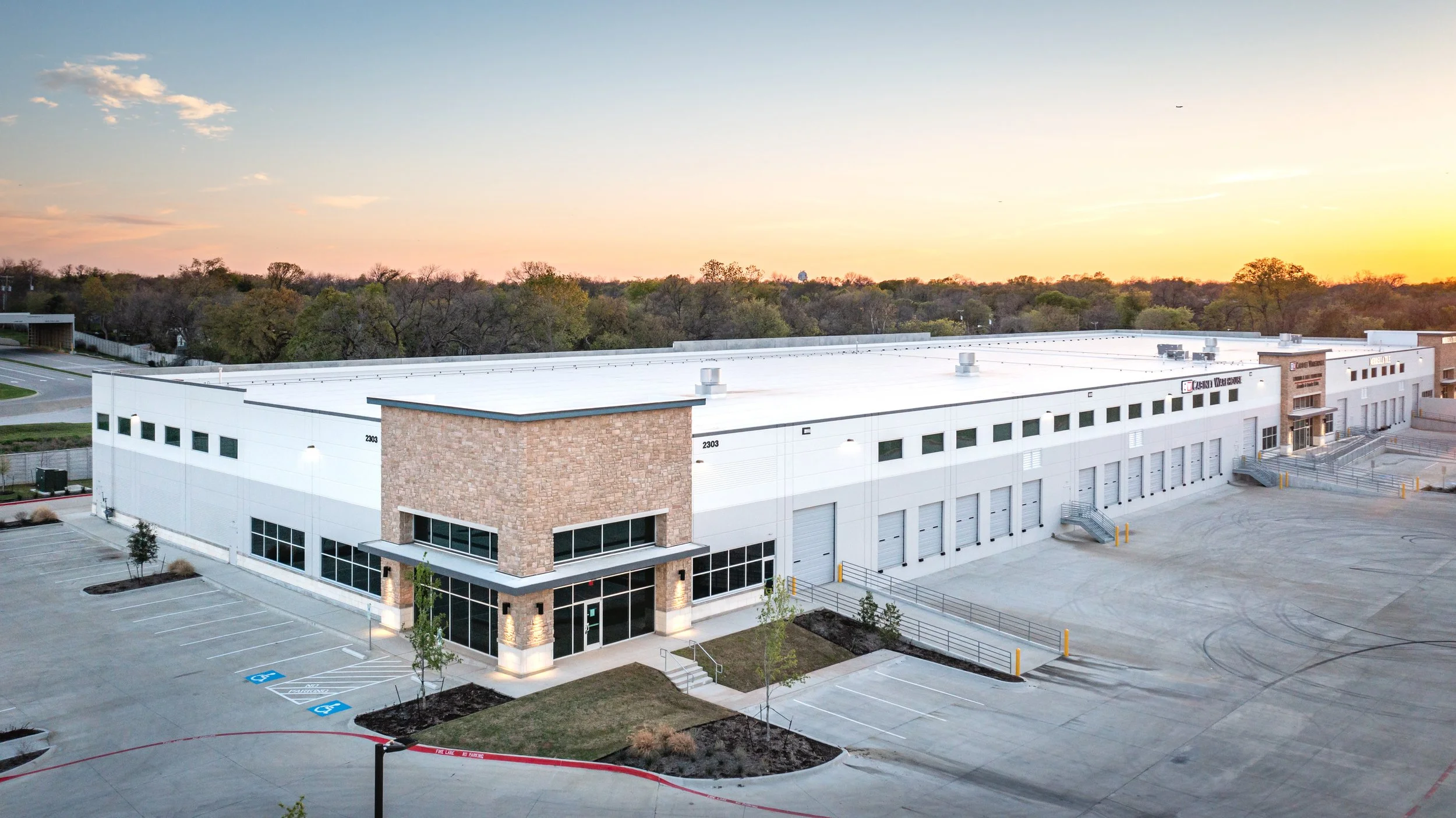 Modern industrial warehouse building with parking lot and trees in the background at sunset.