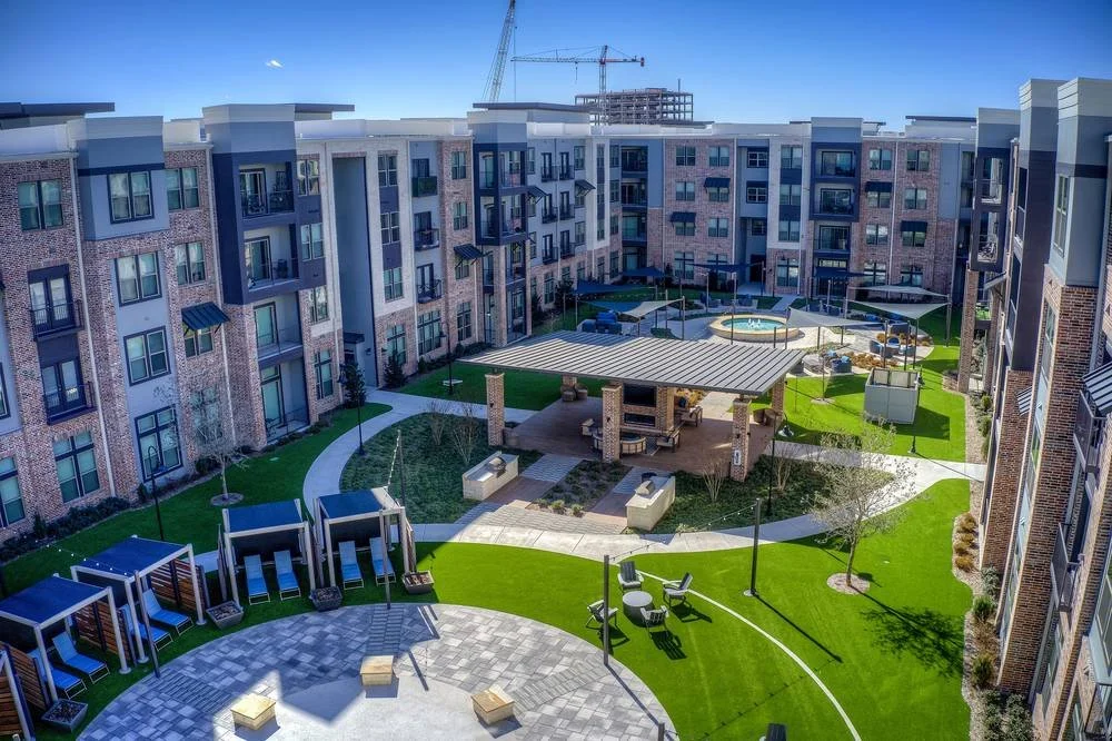 View of a modern apartment complex courtyard with outdoor seating, a covered pavilion, small trees, a swimming pool, and a construction crane in the background.