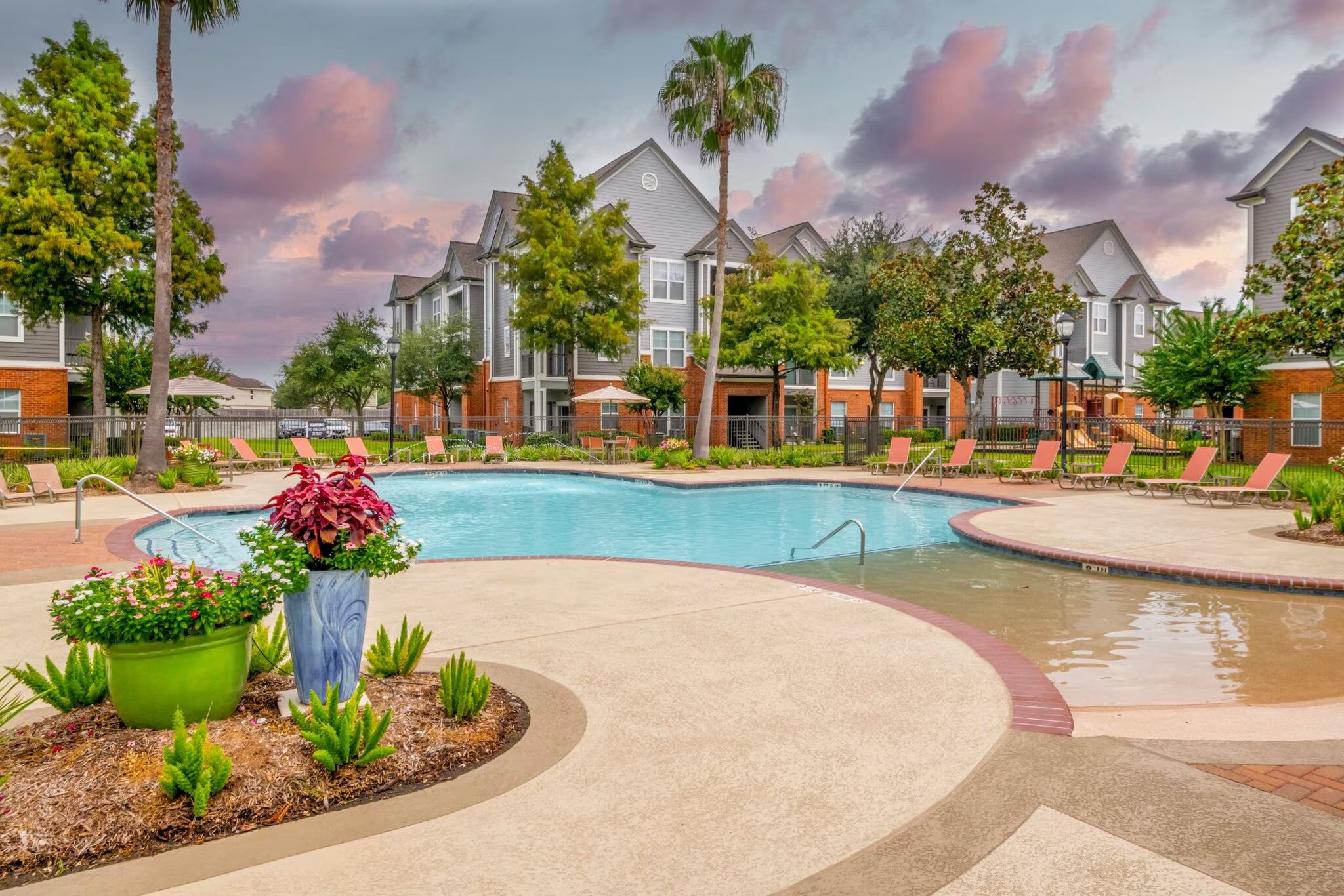 An outdoor swimming pool area in a residential complex, surrounded by lounge chairs, umbrellas, and greenery, with apartment buildings in the background under a cloudy sky.