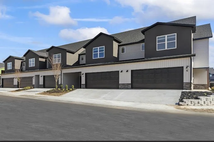 Row of modern townhouses with garages, small trees, sidewalk, and parking area under a partly cloudy sky.