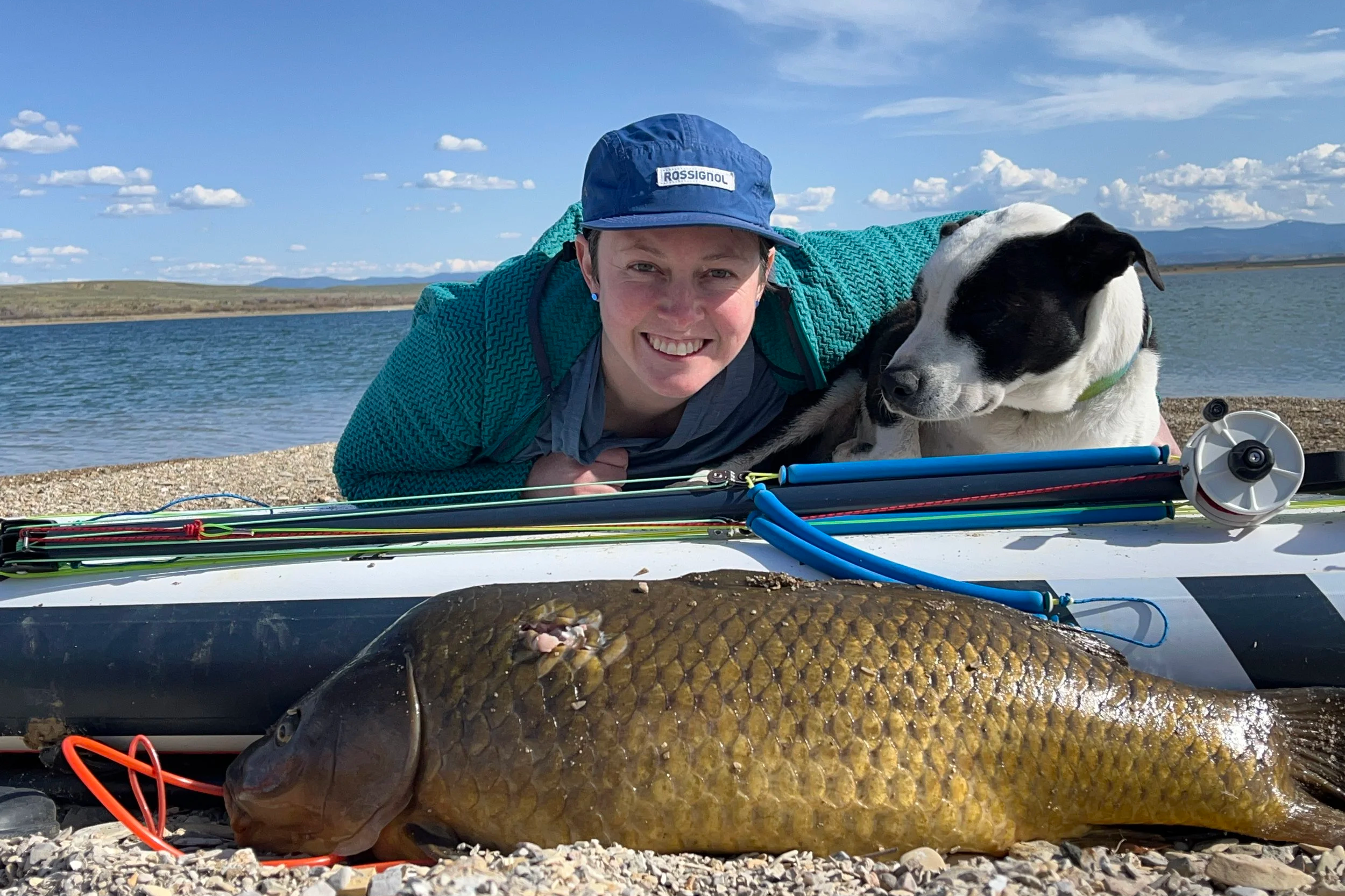 Person lying on the ground next to a large fish with a dog and fishing gear on the shore near a body of water.
