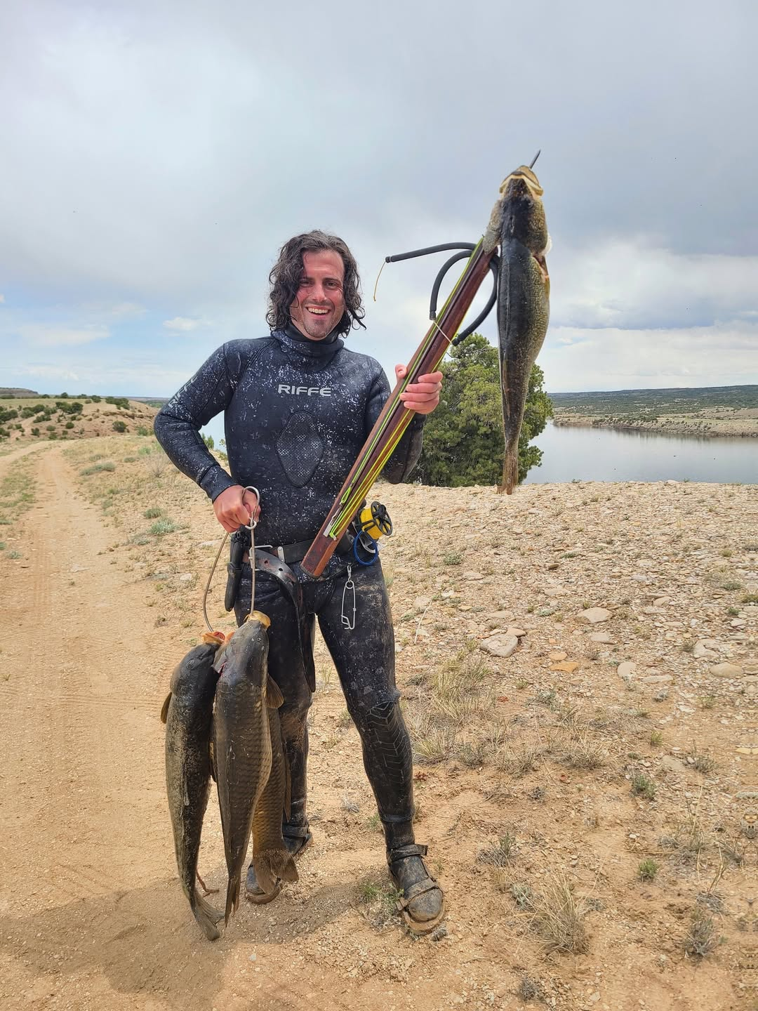 A man standing outdoors on a dirt path holding a fishing spear with a large fish and three smaller fish hanging from his hand, with a river and cloudy sky in the background.