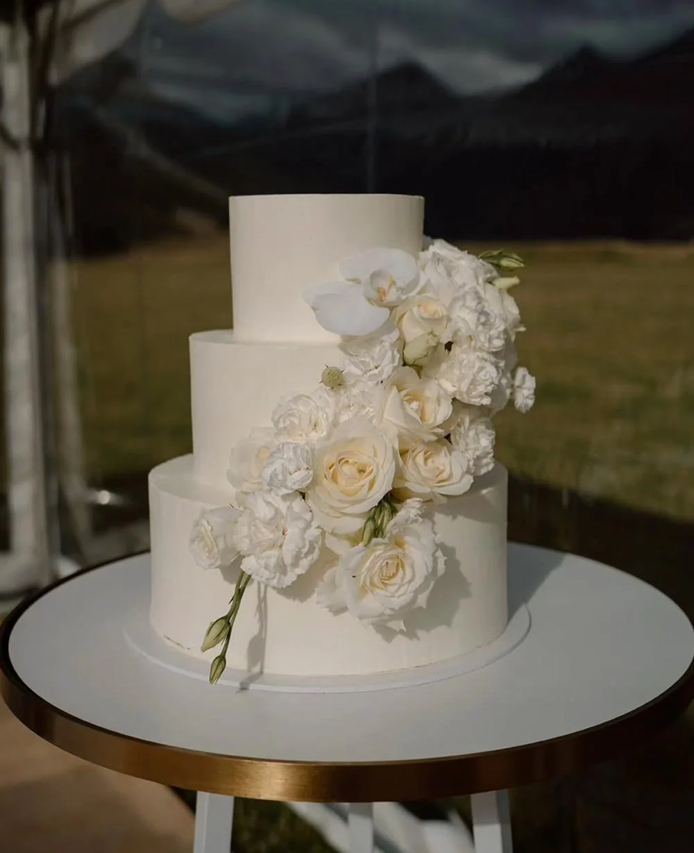 Three-tiered white wedding cake decorated with white orchids, roses, and peonies.