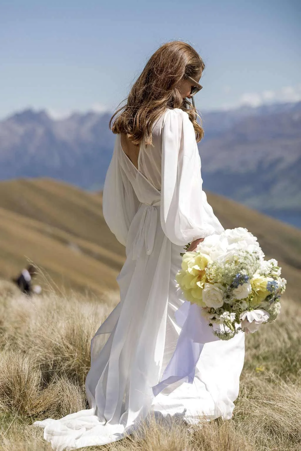 Woman holding a bouquet of white and yellow flowers, standing in a grassy field with mountains in the background.
