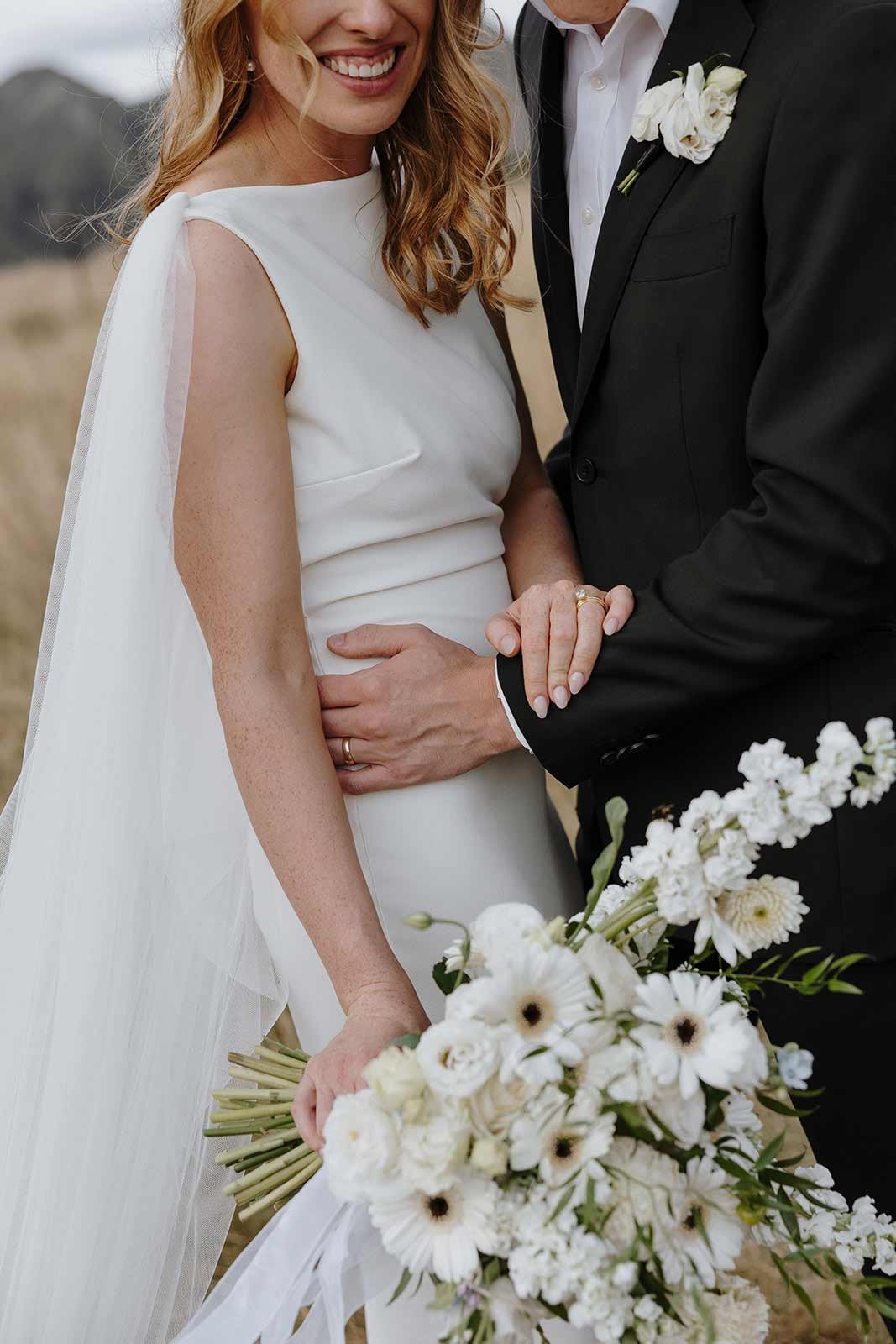 Close-up of a bride in a white dress and a groom in a black suit holding a bouquet of white flowers outdoors.
