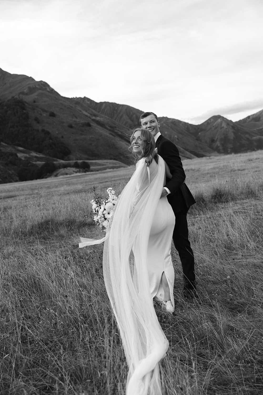 A black-and-white photo of a bride and groom standing in a grassy field with mountains in the background. The bride is holding a bouquet of flowers.