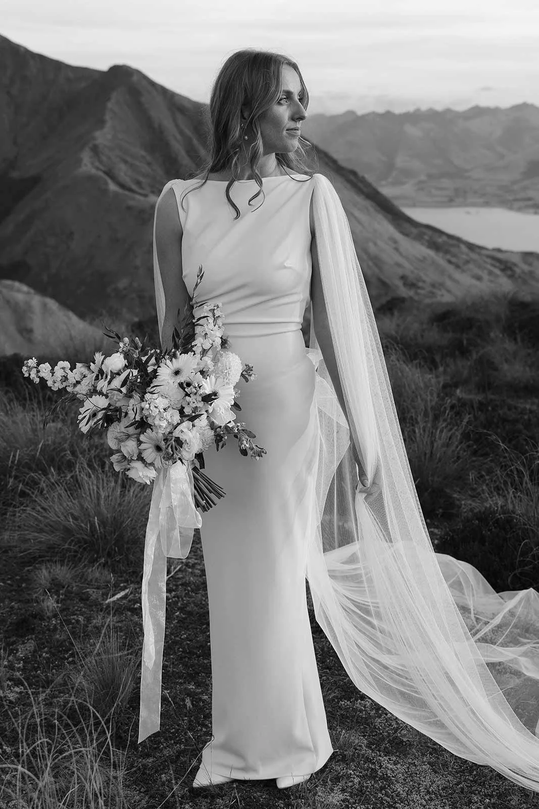 A bride in a wedding dress holding a bouquet of flowers, standing outdoors in a mountainous landscape.