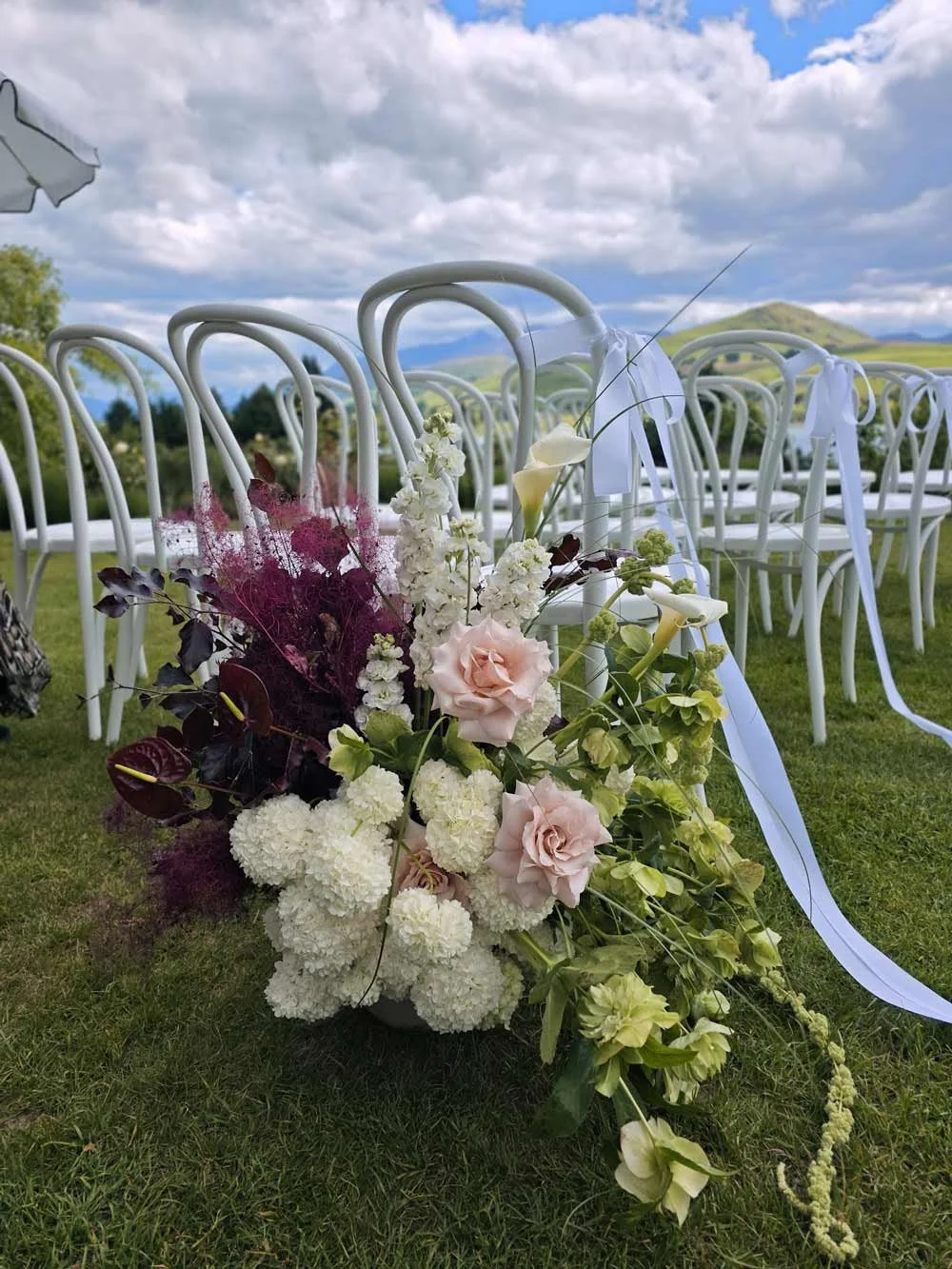 Wedding floral arrangement with white and light pink flowers placed on green grass with white chairs in rows, outdoor setting with hills and cloudy sky in background.